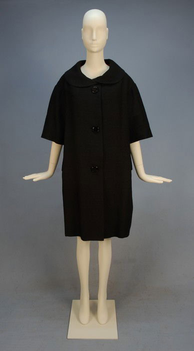 BALENCIAGA COUTURE BLACK WOOL COAT, 1957-1958. Box: BALENCIAGA COUTURE BLACK WOOL COAT, 1957-1958. Boxy cut with elbow length sleeve, wide round collar, 2-piece side panels creating flaps over welt pockets, three oversize composition buttons, back vent