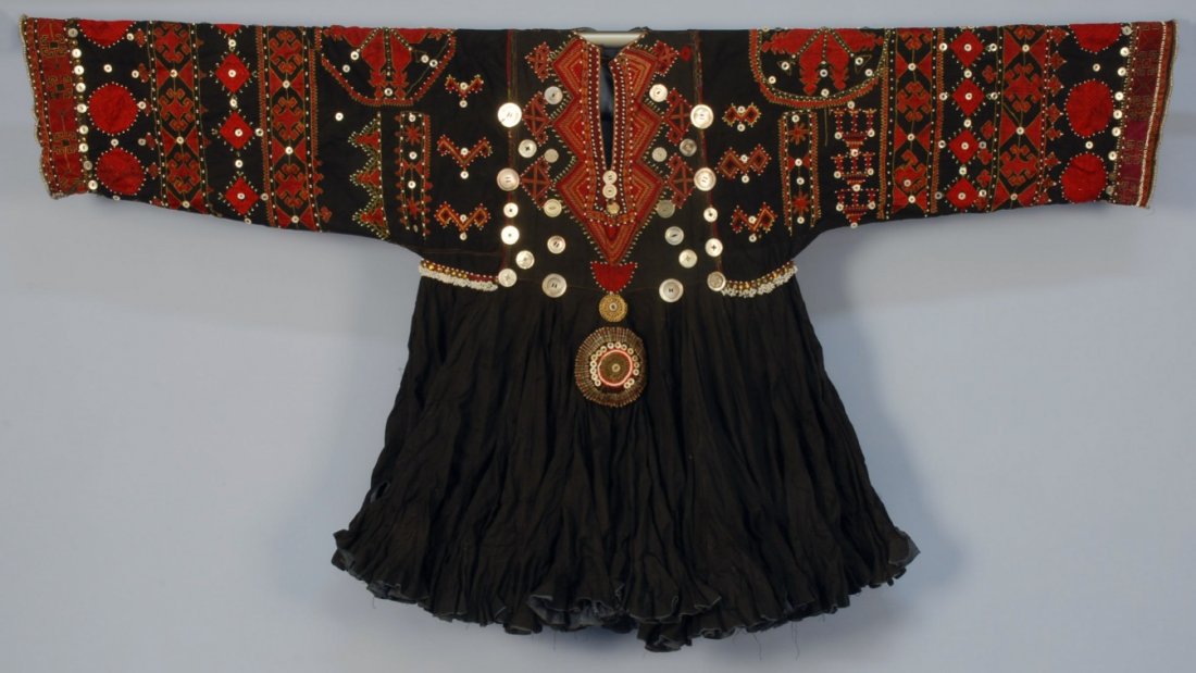 TURKMEN EMBROIDERED DRESS, 20th C.: Long sleeve black cotton with Swat region tribal decoration having embroidery, appliqued beads, mother of pearl and metal buttons, large metal ornament applied below waist, many gored skirt with ruffl