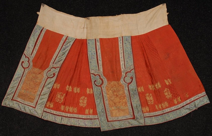CHINESE CEREMONIAL SKIRT, EARLY 20th C: Pleated red silk brocade with appliqued panels of satin stitch embroidery and couched gold metallic thread. In two panels, each 28 1/2 x 39. (Faint stain, loose seams on applique and loose couching th
