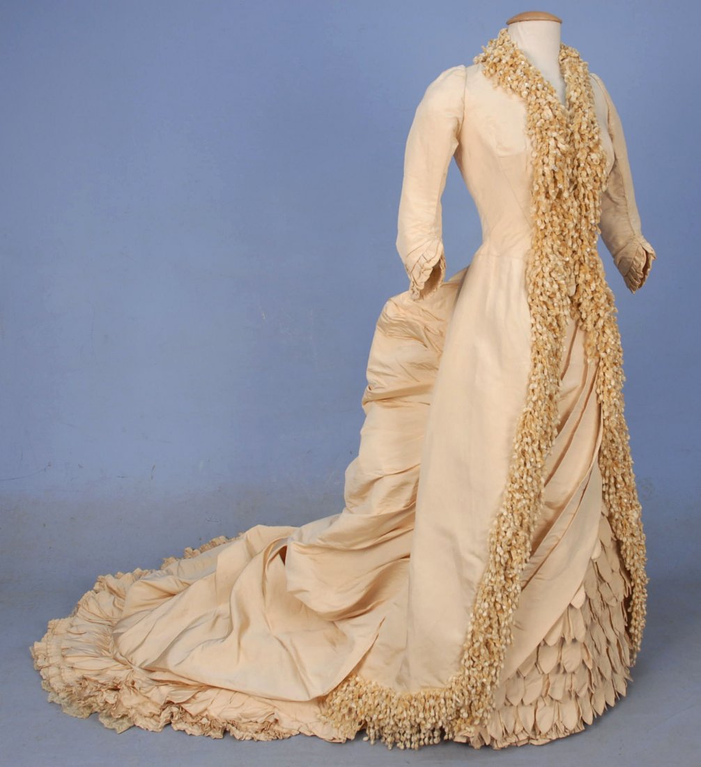 PARIS LABEL WINTER WEDDING GOWN and ACCESSORIES, 1883.: 1-piece ivory silk faille having boned polonaise bodice with 3/4 sleeve trimmed in heavy chenille fringe with white beads. Underskirt draped in angled wide pleats over rows of ruffles comprised of poi