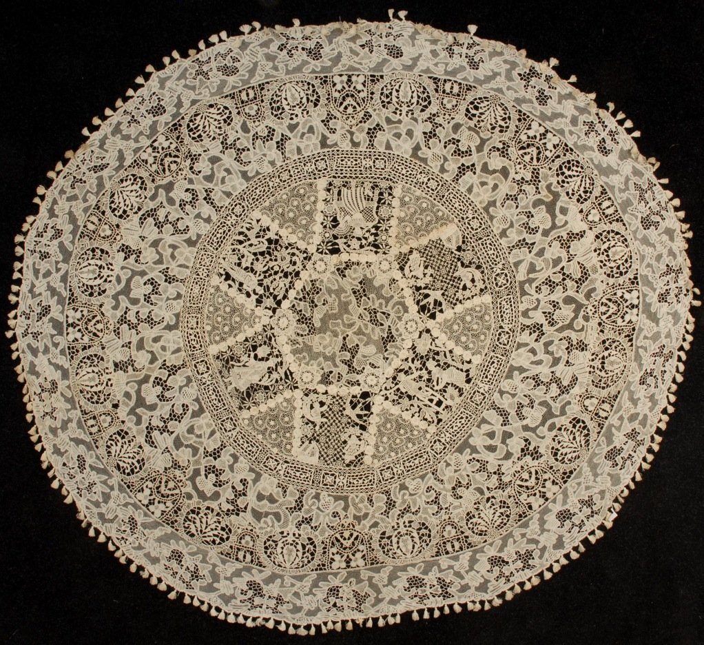ROUND MIXED LACE TABLECLOTH, c. 1915.: Pieced bands of cream needle, bobbin and crochet lace with tasseled border, the interior band having needle lace figures. Diameter 75. (Few broken brides and spots) very good. FIT.