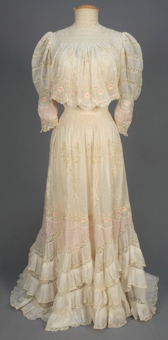 EMBROIDERED TEA GOWN with VALENCIENNES LACE, c. 1899.: White cotton lawn with lace high neck and bands in a pattern of loops and bows, having a band of polychrome embroidered roses at lower bodice, sleeve and skirt, the sleeve and skirt with scalloped ins