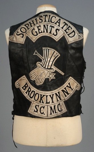 LEATHER MOTORCYCLE CLUB VEST, 20th C.: Black side-lacing style having appliqued "Sophisticated Gents/ Brooklyn N.Y./ S.C. M.C." on back, patches and personalization on front. Protech label. Ch-44, L-25. (Some wear and soiling) good.