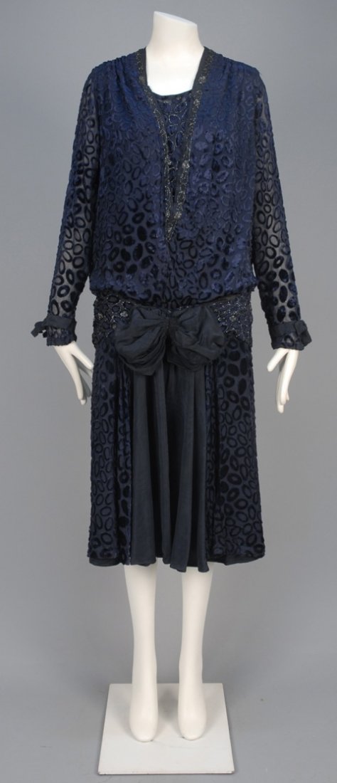 VOIDED VELVET DRESS with BEADED TRIM, 1920s.: Midnight blue velvet cut to chiffon in a pattern of oval rings and spots having long sleeve, beaded V-neck, insert and sash, gathered skirt open over moire silk underskirt with large bow. Label "Black