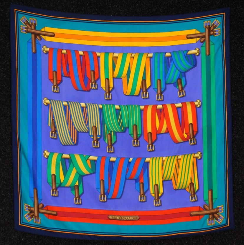 HERMES SANGLES PRINTED SILK SCARF, ISSUE DATE 1985.: Pattern of brightly colored webbing with straps on blue ground. Signed J. Metz. Labeled within design. 35 x 35. (Very faint spot). Together with a Givenchy floral printed silk scarf. 34 x 35. Very goo