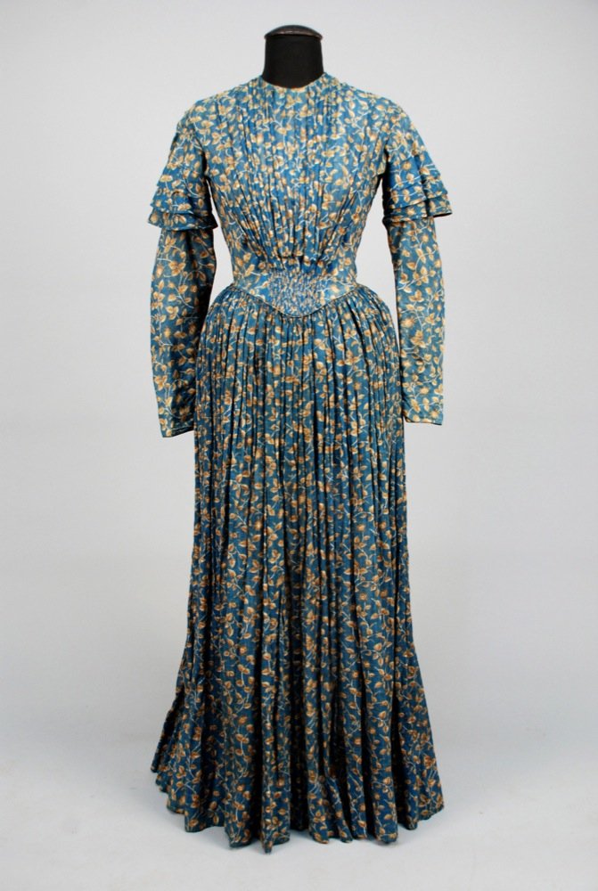 PRINTED WOOL DAY DRESS, 1840s. Blue having woven: PRINTED WOOL DAY DRESS, 1840s. Blue having woven windowpane and printed with leaves and buds in cream and shades of brown, long fitted sleeve capped with triple ruffle, pleated bodice front gathered i