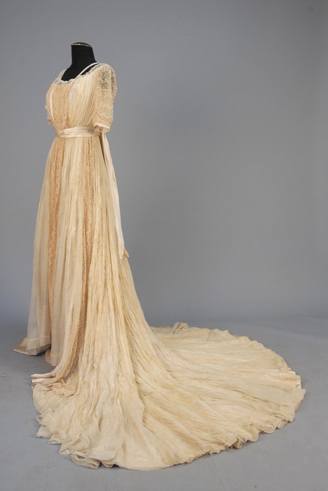 TRAINED EDWARDIAN SILK WEDDING GOWN. Pleated ivory: TRAINED EDWARDIAN SILK WEDDING GOWN. Pleated ivory chiffon over satin, the boned bodice having tulle front panel and short sleeve with serpentine vines and berries, satin trim and sash with streamers,