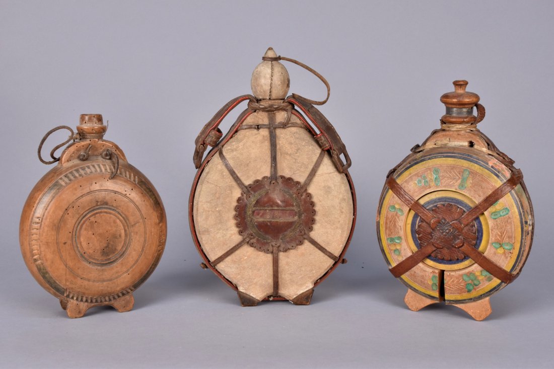 THREE HUNGARIAN FOOTED WOOD FLASKS, 1880s and EARLY: One carved in a pattern of circles with hide cord handle, 6 1/2 x 9 1/2, (lacking stopper, worm holes, cracked. One covered in hide decorated with embroidered leather including "1887", belted around t