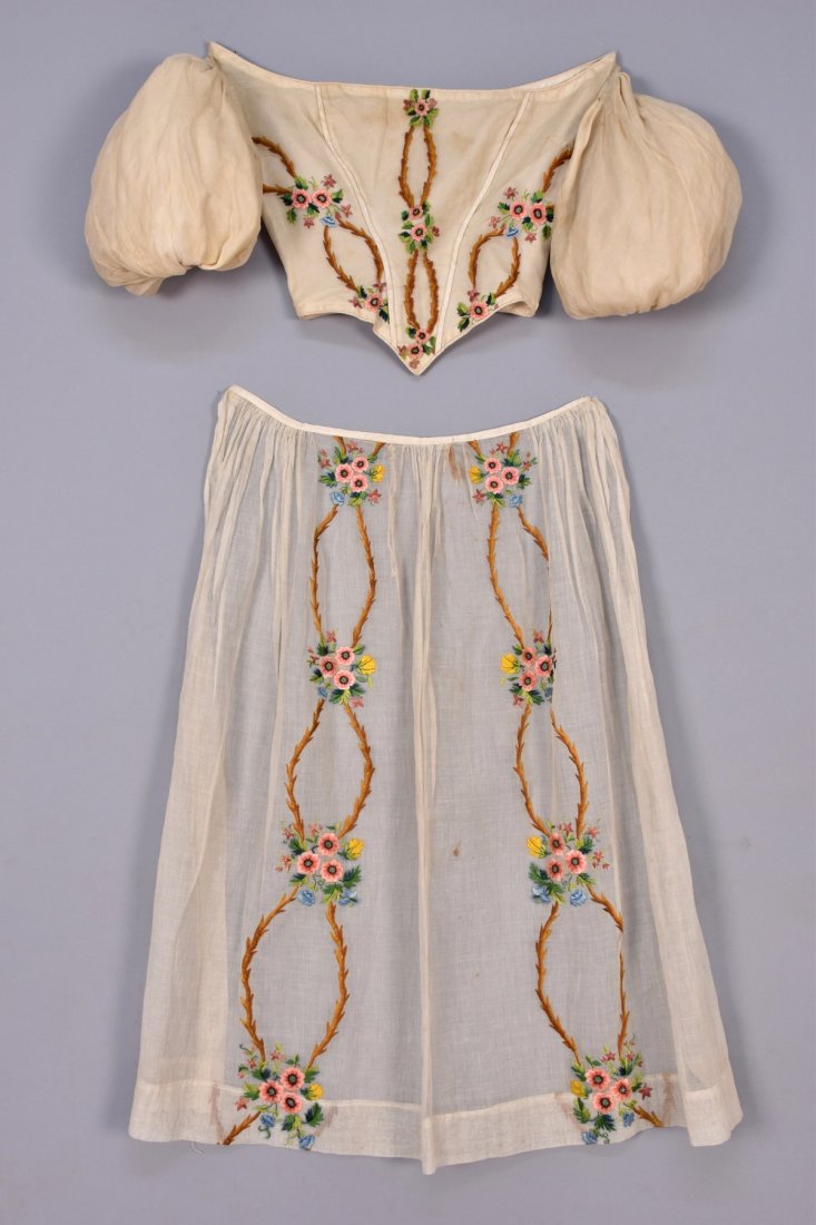 EMBROIDERED BODICE and APRON, 1830s.: White cotton batiste bodice having short melon sleeve and front point with silk piping decorated in polychrome satin stitch flowering branchwork, silk lining, matching unlined apron with cord tie. B-3