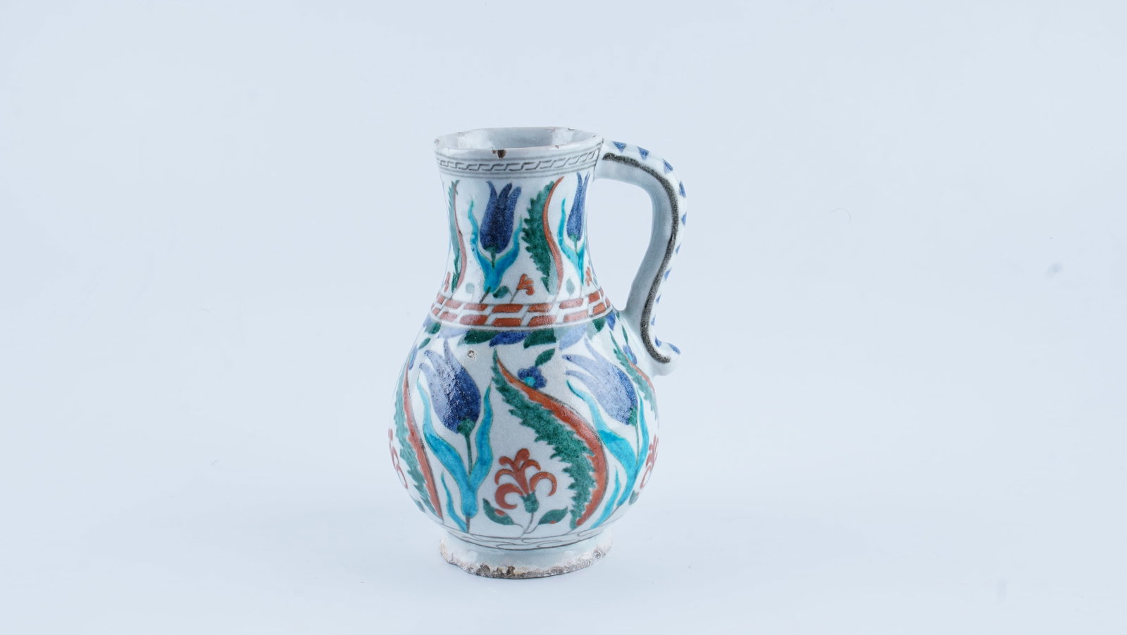 AN OTTOMAN IZNIK POLYCHROME POTTERY JUG WITH TULIP DECORATION, 17TH CENTURY (CIRCA 1610–1630): A fine example of early 17th-century Iznik polychrome pottery, this jug reflects the naturalistic floral style associated with Ottoman ceramic production of the period. Potted in fine white fritware, 