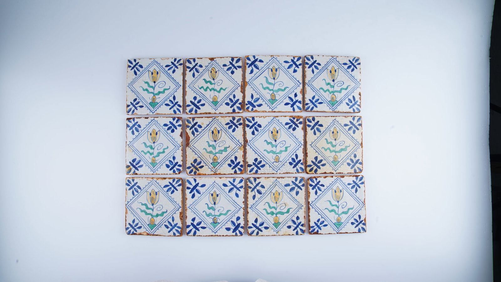 FINE SET OF TWELVE EUROPEAN POLYCHROME TILES IN THE DELFT MANNER, 17TH/18TH CENTURY STYLE: A fine set of twelve polychrome pottery tiles decorated in the Delft manner, each centered with a stylized floral motif enclosed within a diamond-shaped panel. Executed in cobalt blue, yellow, green, 