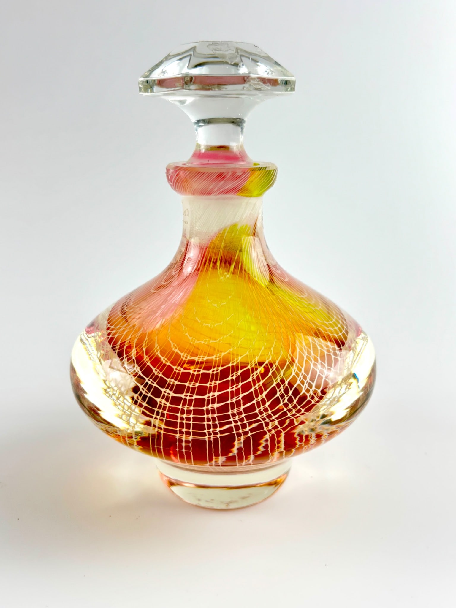 Art Glass Perfume Bottle : Art glass decanter featuring a bulbous body with multicolored swirled patterns in pink, yellow, and orange, overlaid with a white linear net-like design. The glass stopper is clear with a faceted top 
