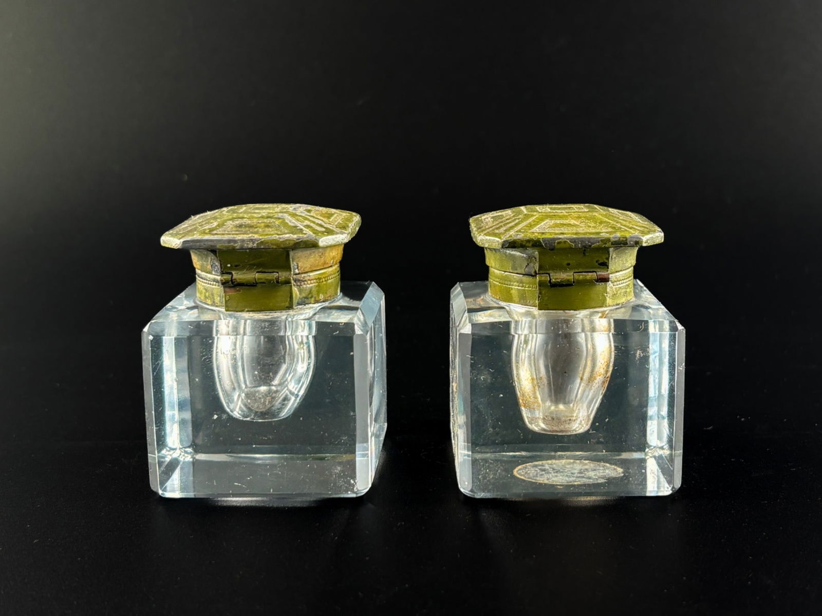 Square Crystal Ink Wells with Octagonal Hinged Metal Lids: Square crystal ink wells featuring leaded glass reservoirs and hinged metal lids with a greenish patina and geometric design. Lids exhibit wear consistent with use, including surface marks and minor c