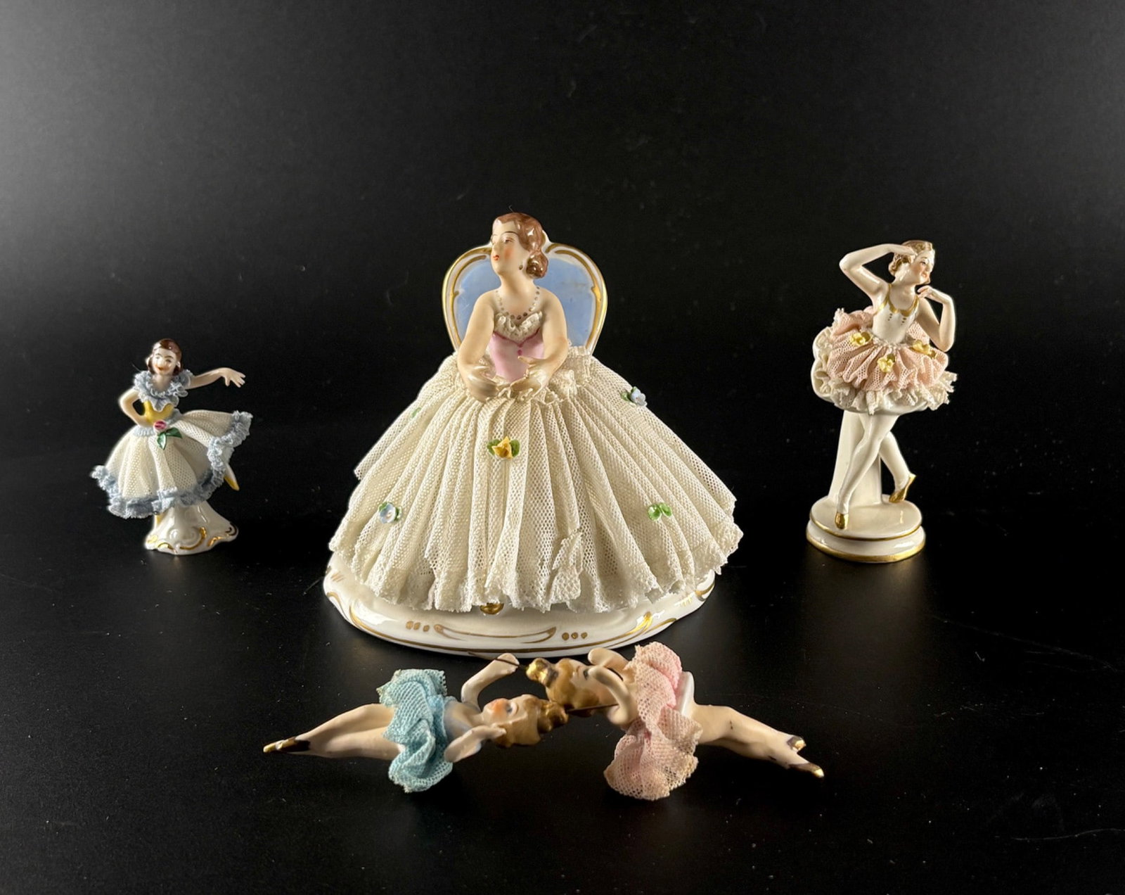 German Porcelain Lace Figural Ballerinas : German porcelain figurines featuring ballerinas with handmade lace skirts and hand-painted details. Includes four standing figurines, one seated figurine with a blue-backed chair marked ‘Made in
