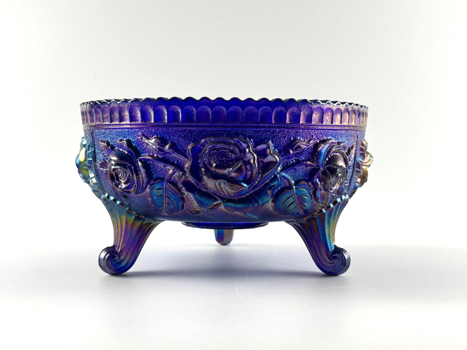 Fenton Carnival Glass Imperial Blue Lustre Three-Toed Rose Bowl: Fenton carnival glass bowl in imperial blue lustre finish featuring a raised rose pattern on the exterior and a ruffled edge. Supported by three scrolled legs with detailed accents. Interior shows sub