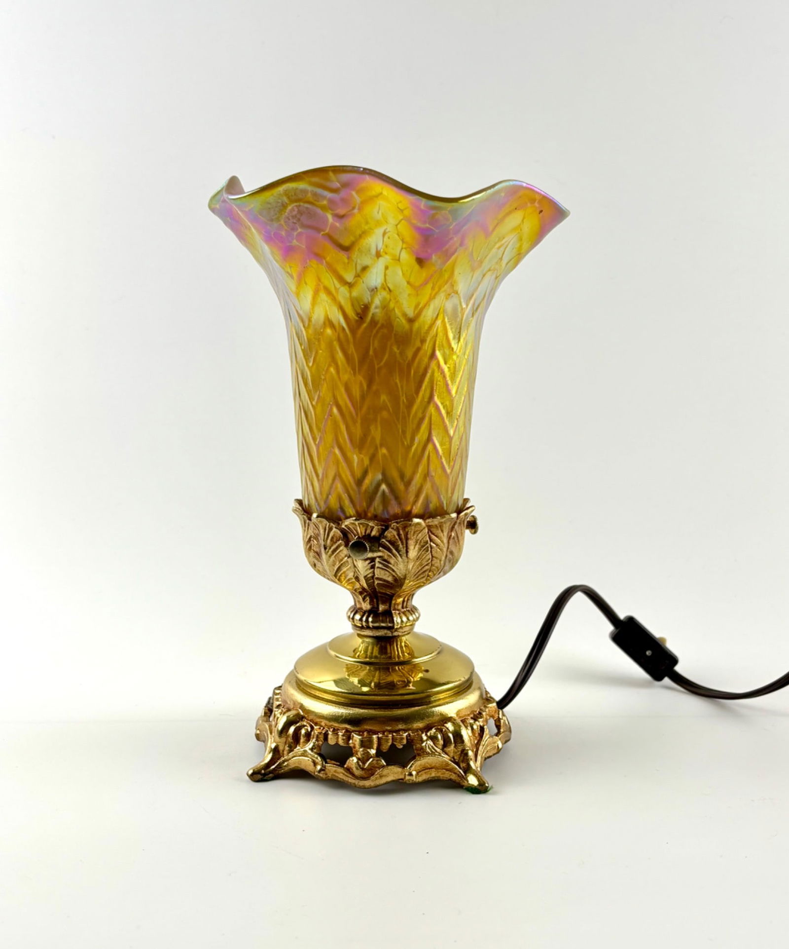 Loetz-Attributed Iridescent Art Glass Lamp with Ornate Gilt Base: Attributed to Loetz Austria, this small electric lamp features an iridescent art glass shade with a chevron design and gold, pink, and purple hues. The flared shade is supported by a gilt metal base w