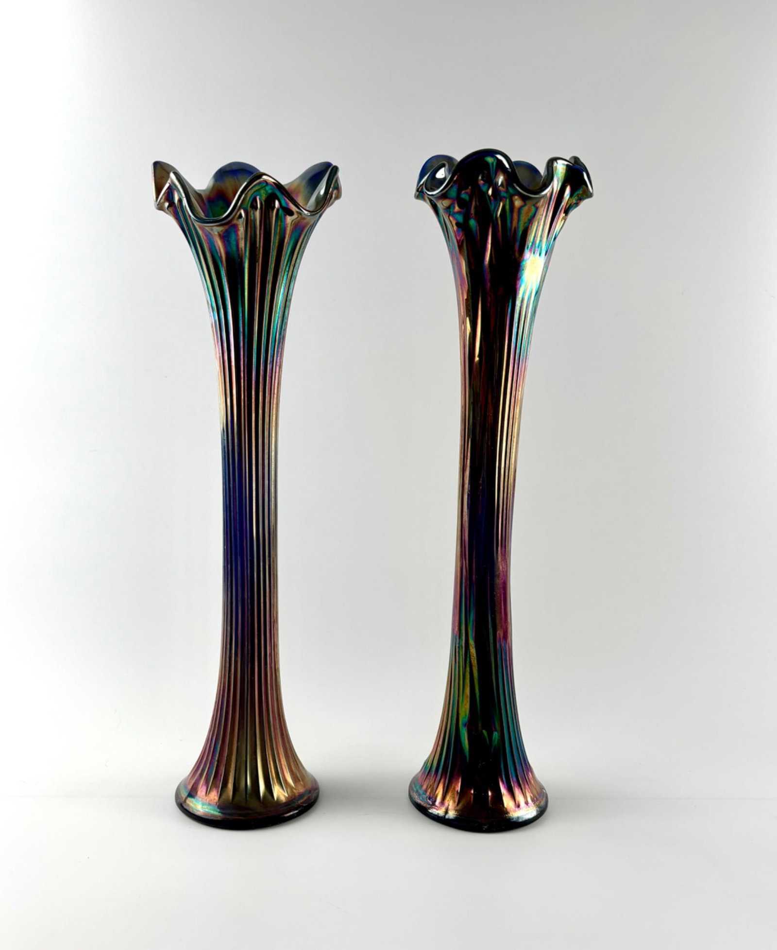 Fenton Carnival Glass Tall Fluted Vases: Fenton Carnival Glass vases featuring a tall, elegant design with fluted bodies and ruffled tops. Exhibiting a vibrant iridescent finish with multicolor highlights. Bases have a decorative pressed sta