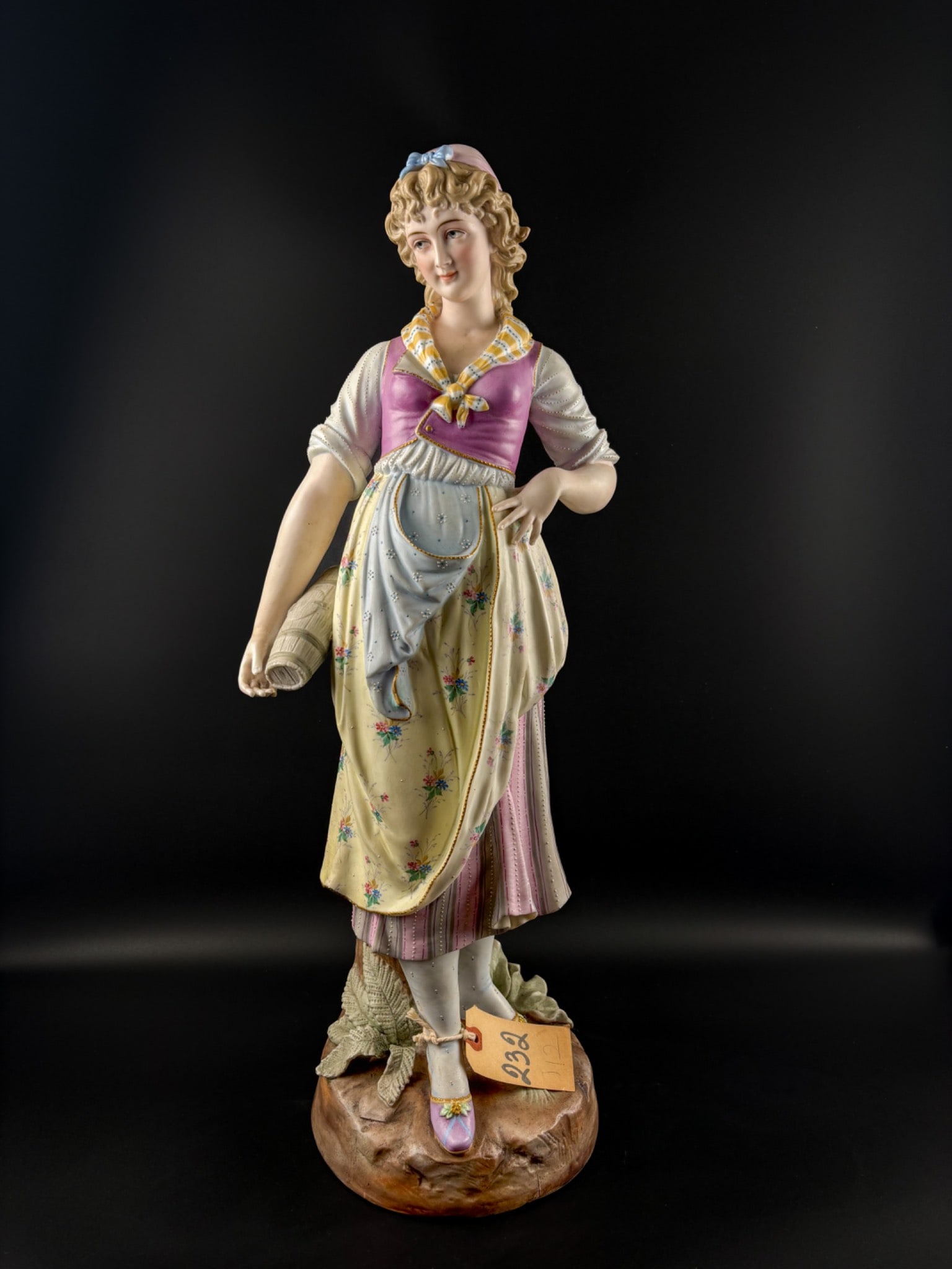 German Bisque Porcelain Figurine (HC, 19th Century): Porcelain statue of a standing woman in traditional attire holding a scroll. Features a hand-painted floral-patterned dress with pastel accents and detailed textured embellishments on the garments. Cr