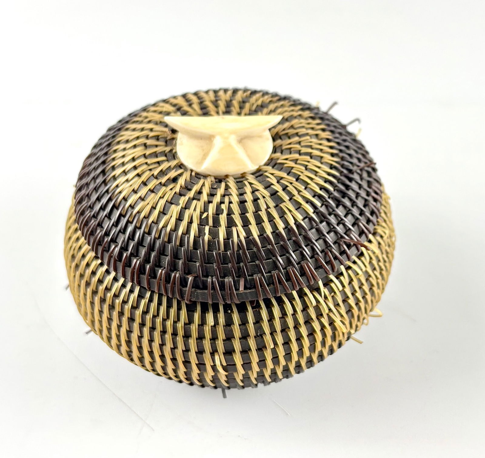 Alaskan Inupiat Baleen Lidded Basket : A lidded handwoven seagrass and baleen basket with dark and light woven materials creating a two-tone design. The lid features a carved ornament in the shape of a whale. The base of the basket is insc