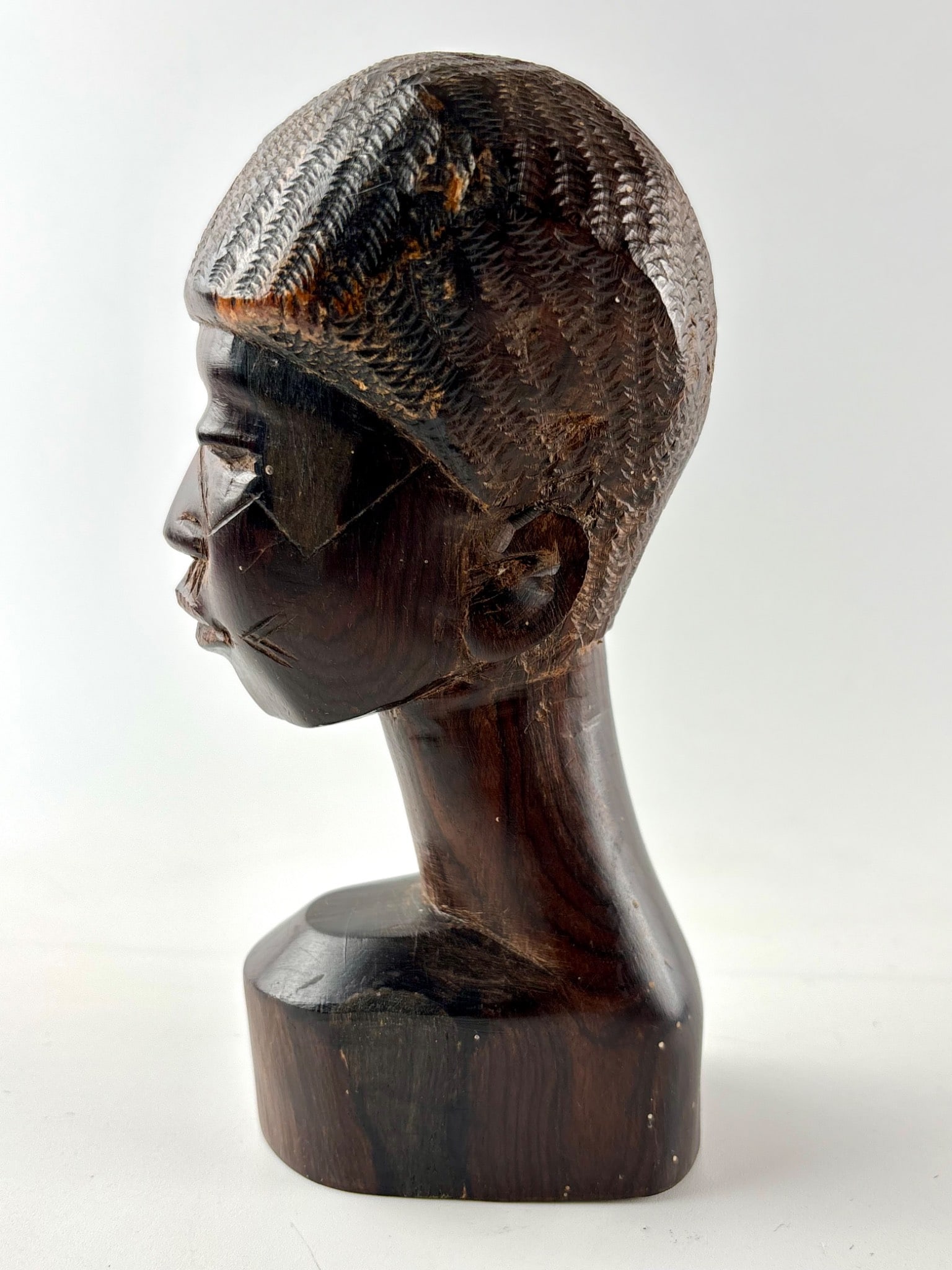 African Hand-Carved Wood Bust with Stylized Facial Features: Hand-carved wooden sculpture depicting a bust with stylized facial features, including scarification details on the cheeks. The figure features a textured hairstyle and is polished, showcasing the nat