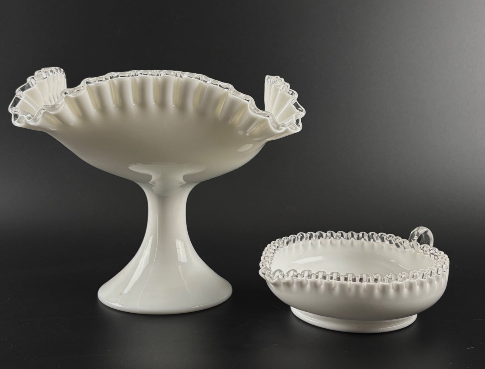 Two White Milk Glass Pieces with Clear Ruffled Edges: Pedestal Bowl and Handled Dish: A white milk glass pedestal bowl and a handled dish, each with clear ruffled edges. The pedestal bowl features a square, flared design with scalloped detailing and a round base. The handled dish is ci