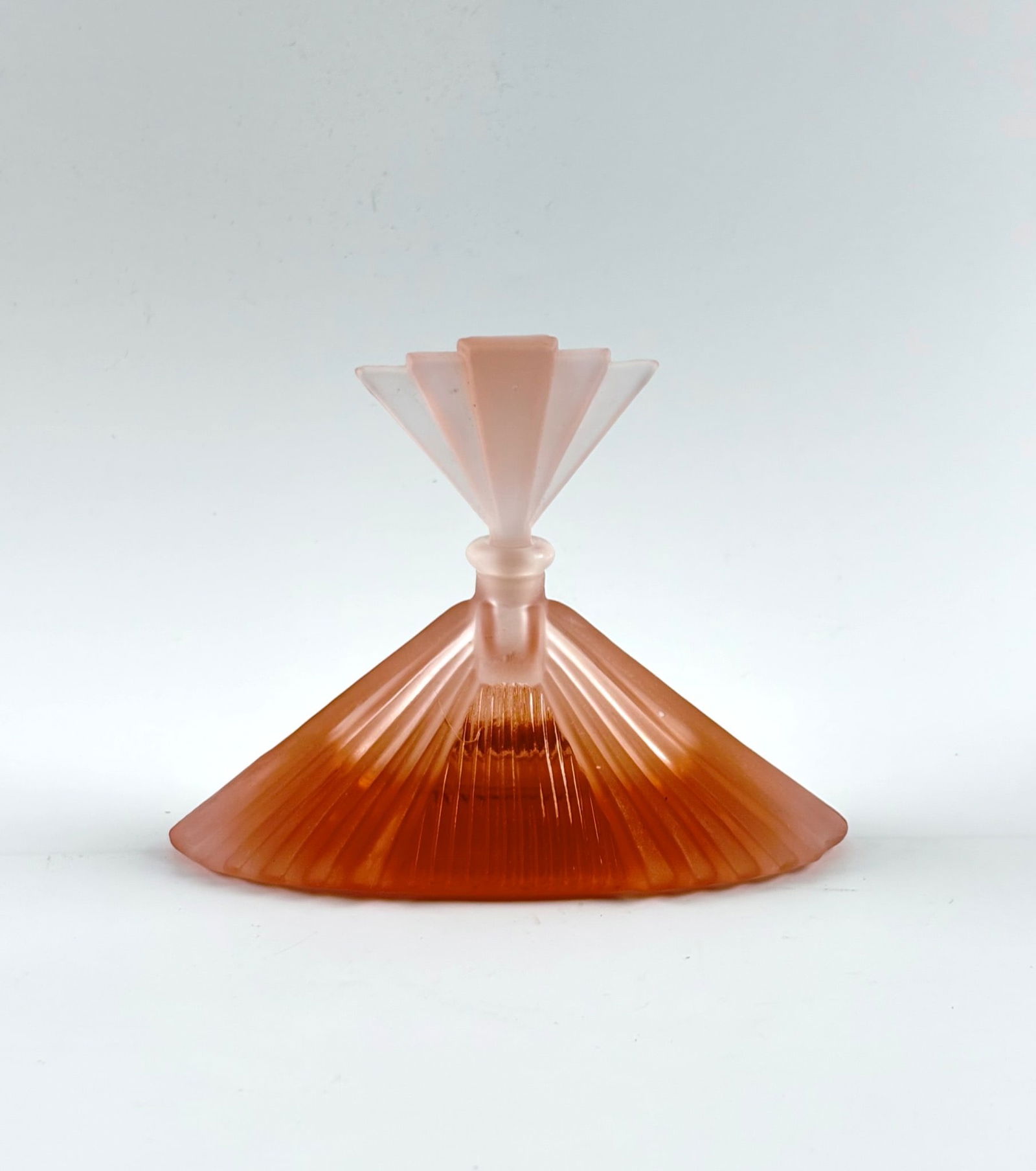 Vintage Art Deco Frosted Glass Perfume Bottle with Fan Stopper: Vintage Art Deco frosted glass perfume bottle in a warm pink hue, featuring a geometric, ridged design motif. The fan shaped stopper complements the angular aesthetic. The stopper has a visible chip,