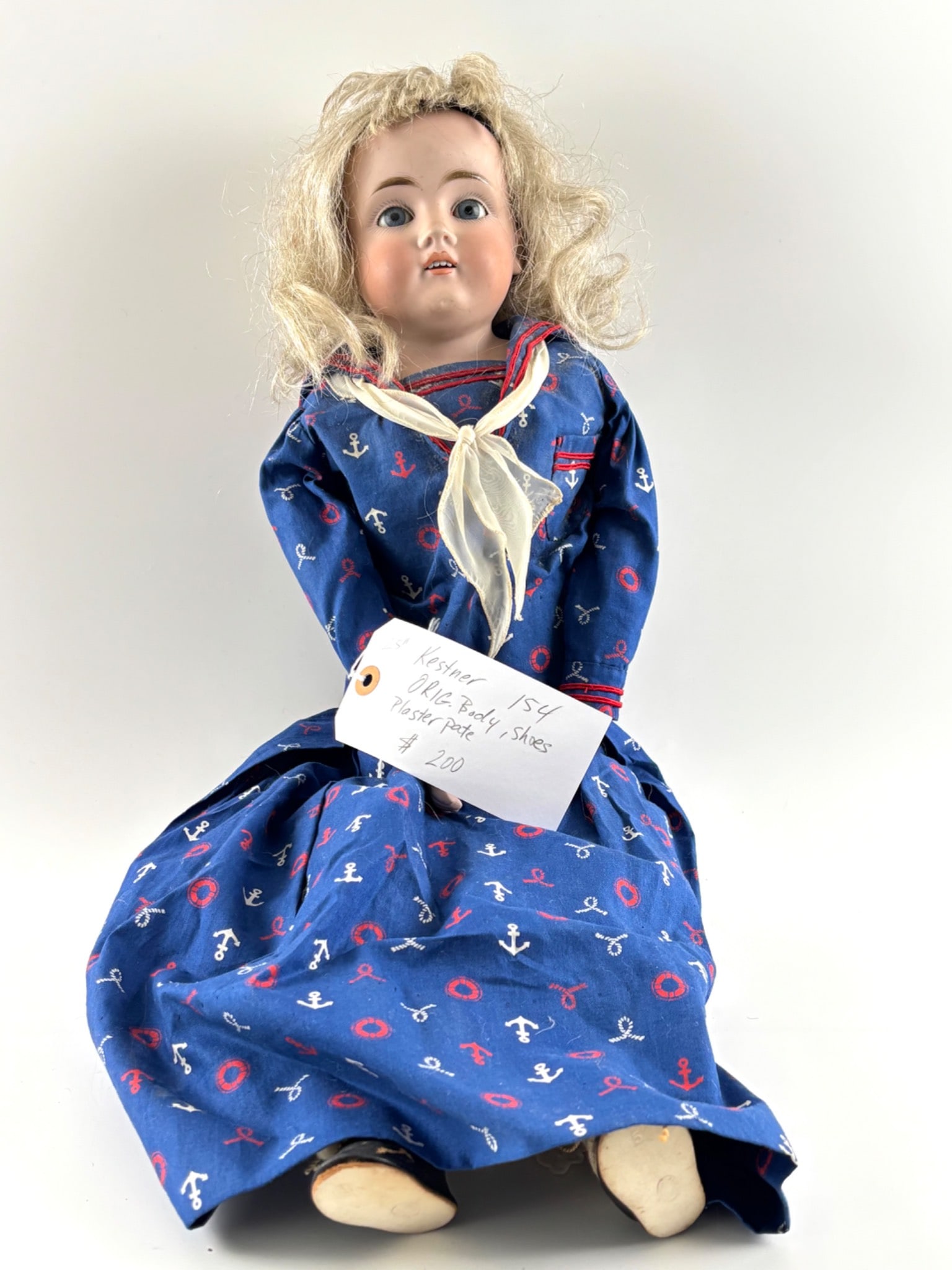25 Inch German Kestner No. 154 Bisque Doll: 25 inch bisque doll marked on the reverse of the head. Features blue sleep eyes, open mouth with teeth, blonde wig, and original body, as indicated on the tag. Dressed in a blue nautical themed dress
