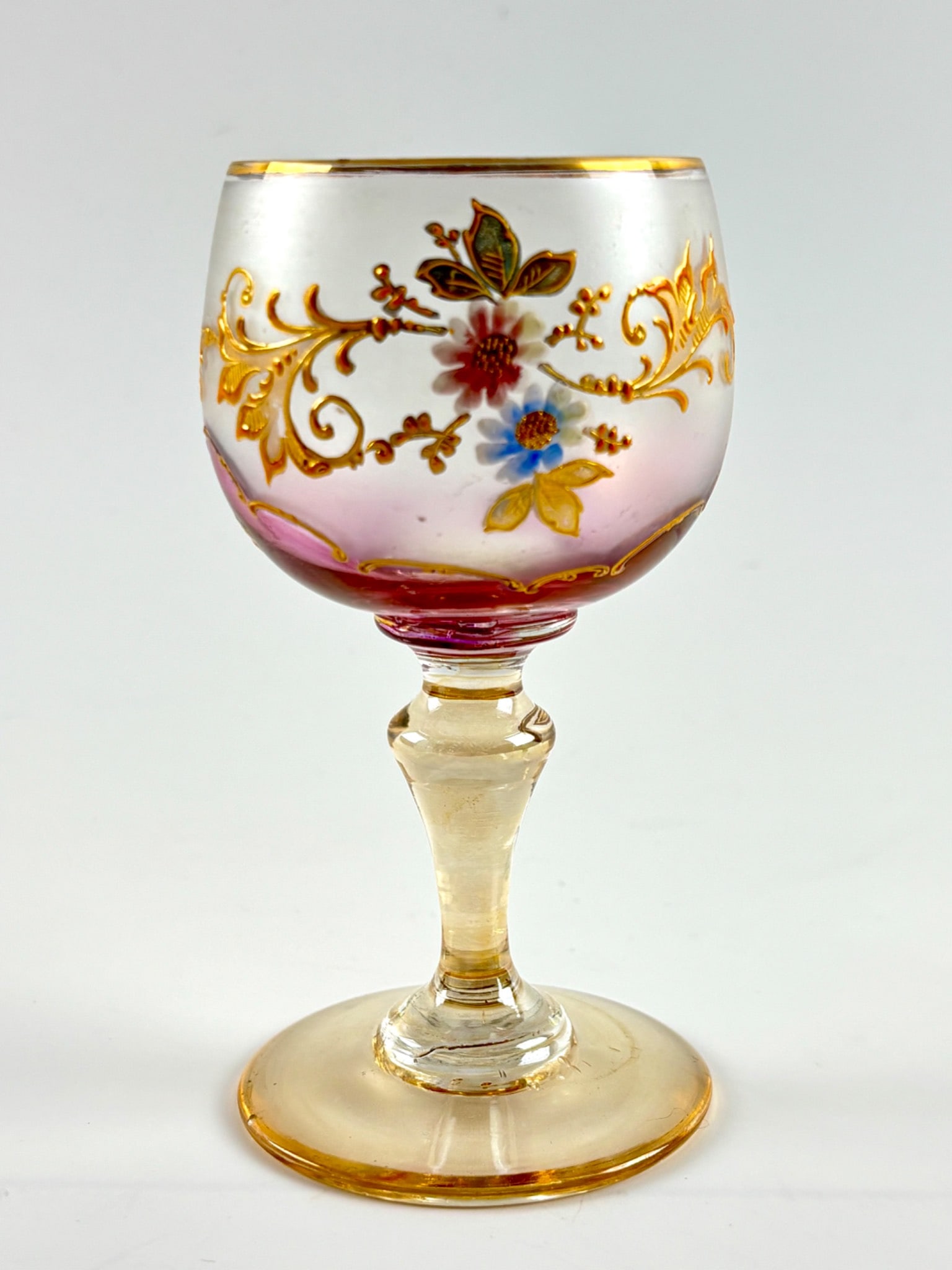 Victorian Hand Blown Cordial with Painted Floral Motif: Victorian era hand blown miniature goblet featuring an enameled floral motif with gold accents on a pink and clear glass body. The goblet showcases intricate details such as scrolling gold patterns an