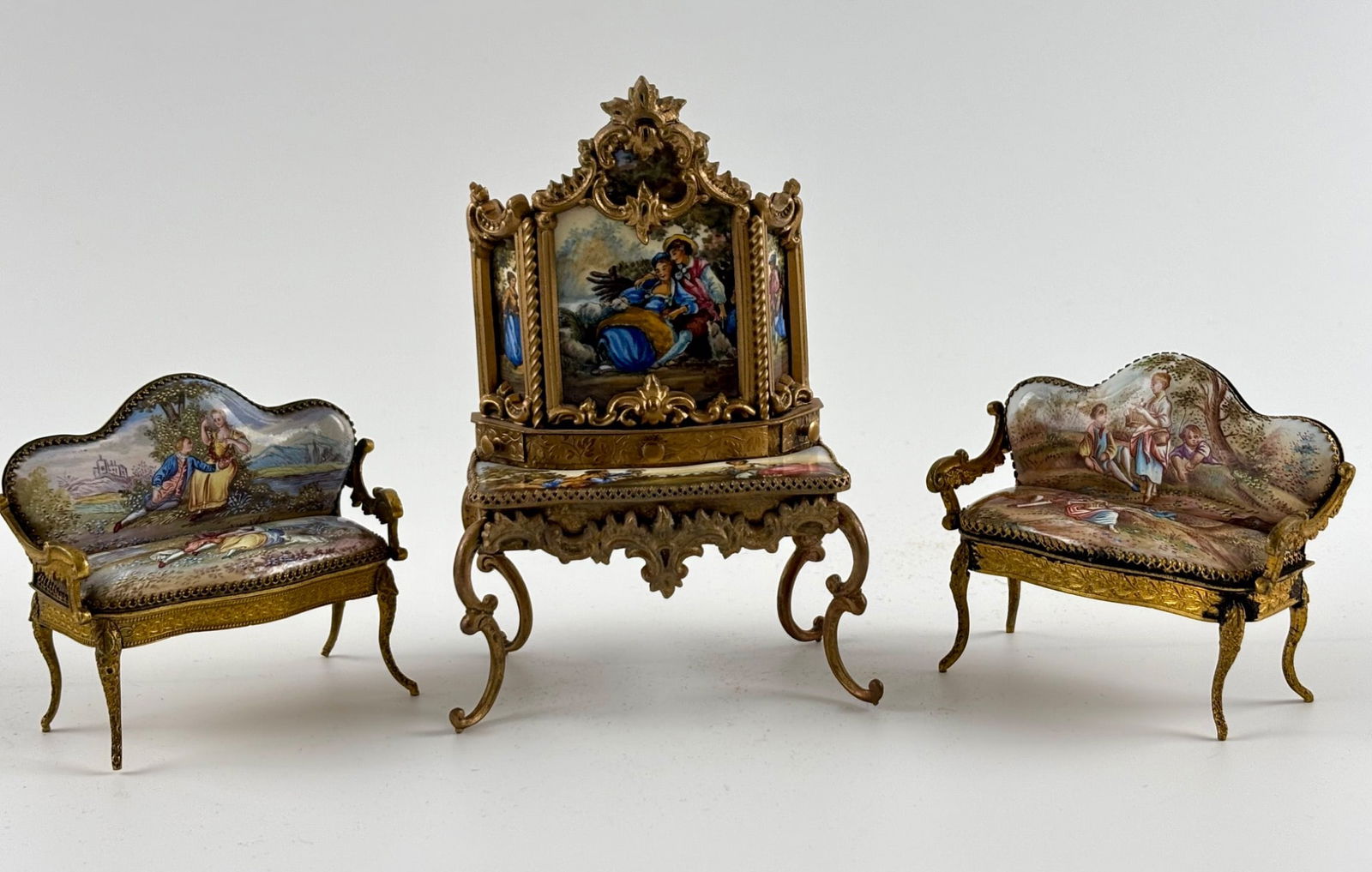 Austrian Gilt Metal and Hand-Painted Enamel Miniature Furniture: A three piece set of Austrian miniature furniture made of gilt metal with hand painted enamel panels. The set includes two settees, and a vanity, each ornately decorated with floral motifs and pastora