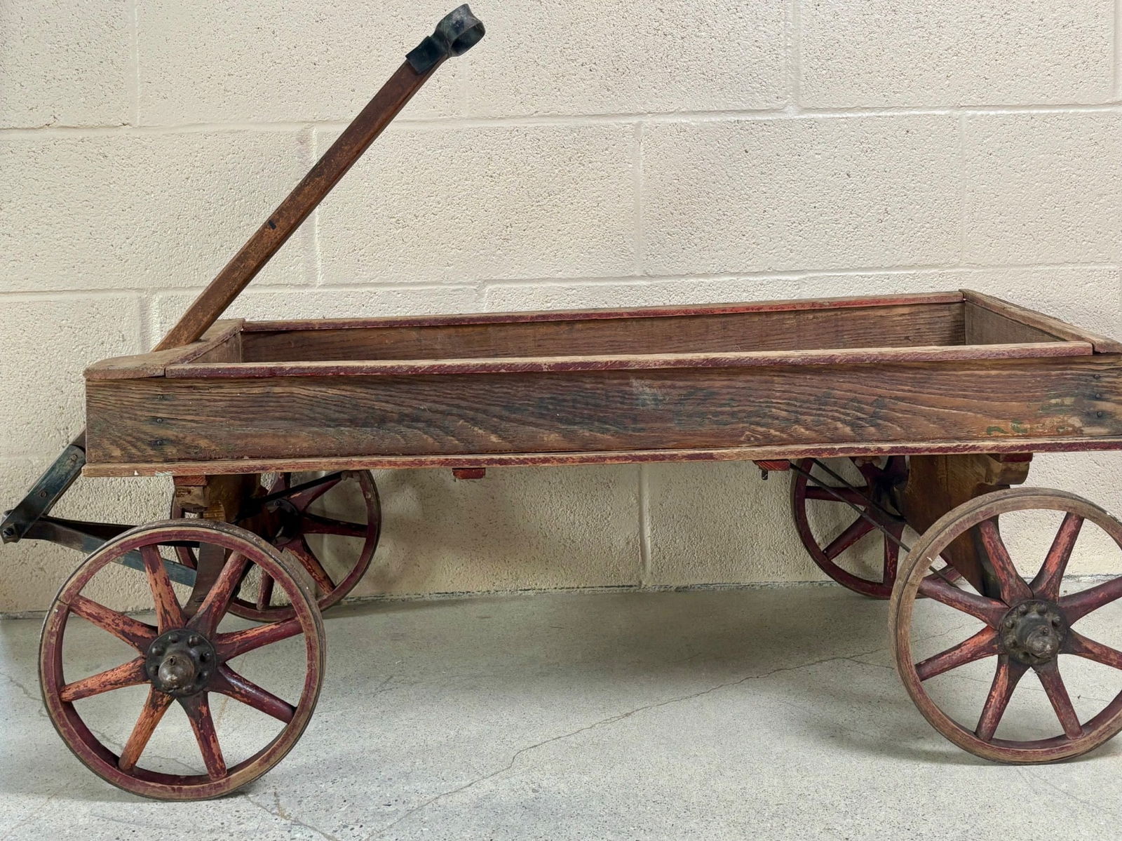 Wooden Pioneer Coaster Wagon with Red Spoked Wheels: Wooden Pioneer Coaster wagon featuring a rectangular frame with a pull handle affixed to the front axle for steering. The wagon is constructed from slatted wood, displaying natural wear, and is affixe