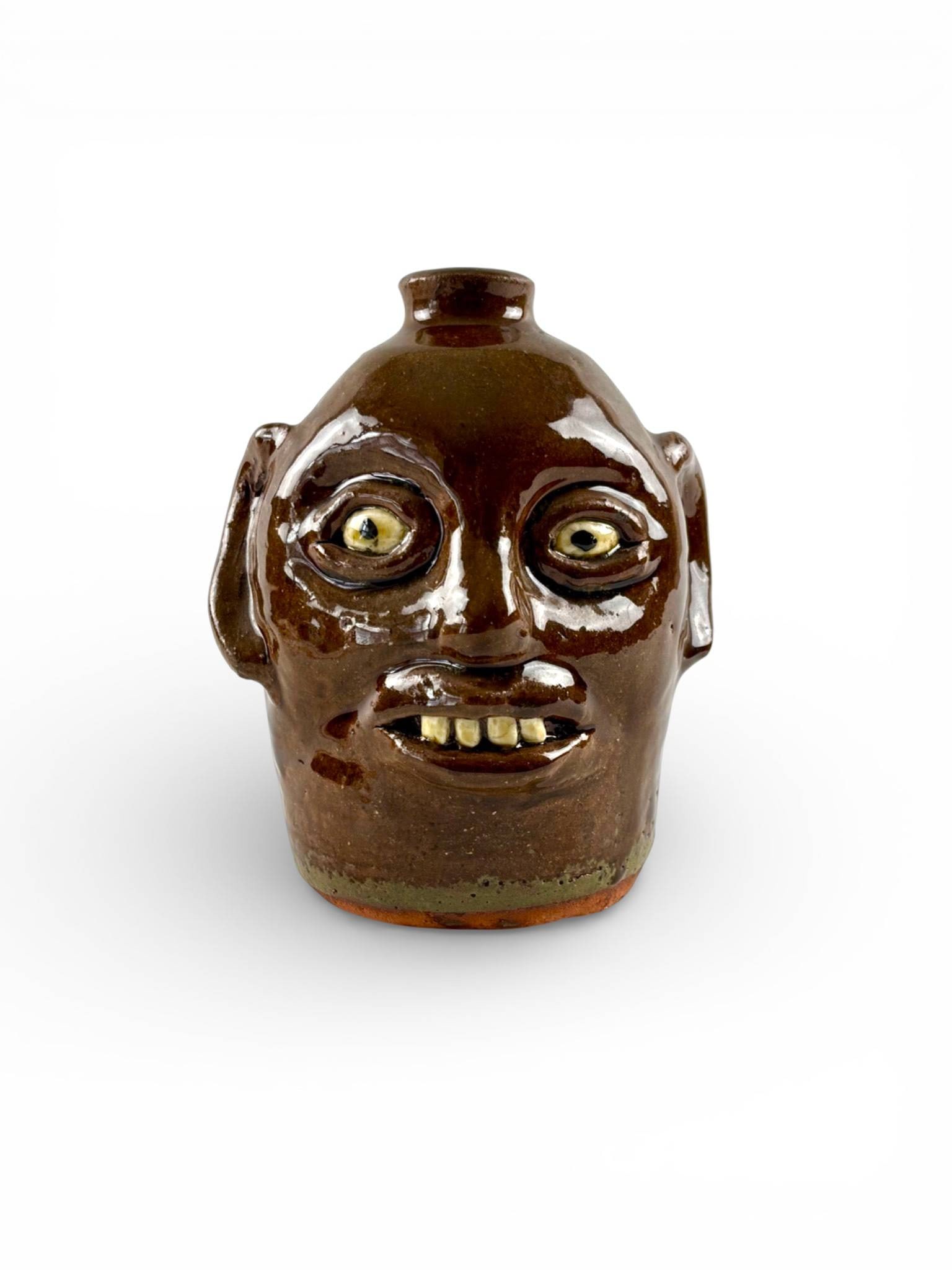 Reggie Meaders Signed Ceramic Face Jug: Reggie Meaders ceramic face jug featuring a brown glaze with applied facial features, including prominent eyes, ears, a nose, and teeth. The jug has a single strap handle and a narrow spout. The