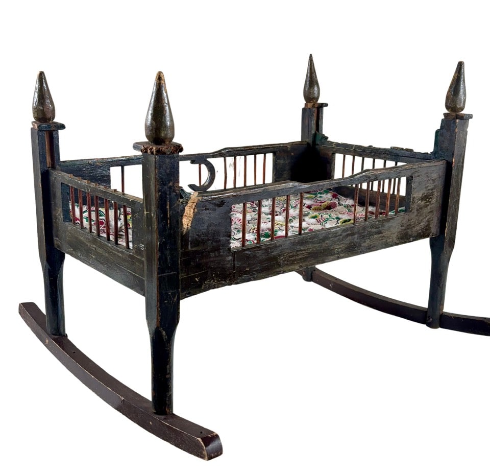 Earlt 19th Century Folk Art Rocking Cradle: Wooden rocking cradle featuring a spindle design along the panels and pointed finials on the posts. The structure includes carved details and a curved rocker base. The cradle is lined with a floral