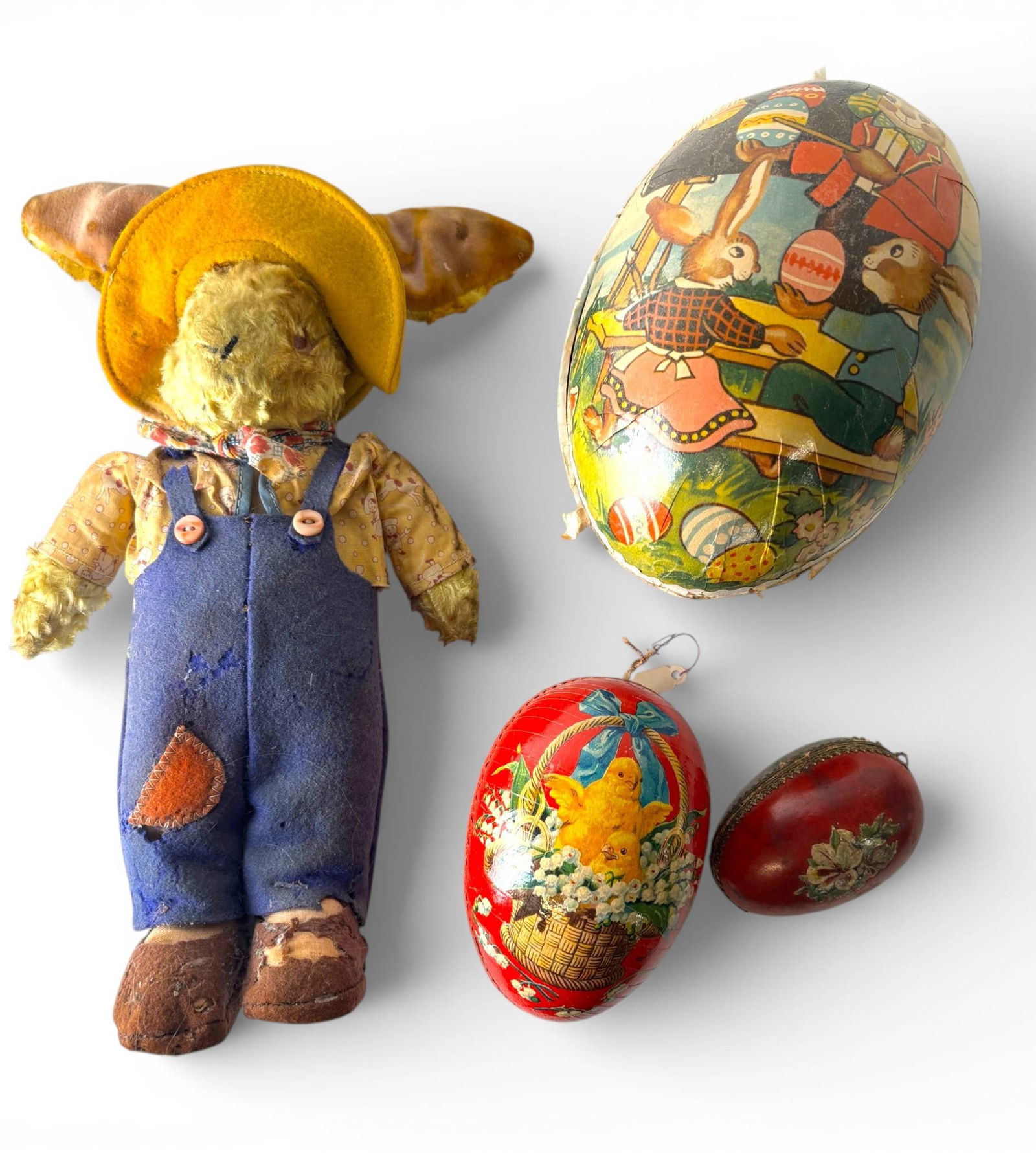 Antique Straw Rabbit and Paper Mache Easter Egg Lot: A vintage plush rabbit toy with yellow felt hat, patterned fabric shirt, and blue overalls, showing wear with patches and frayed detailing. Accompanied by three paper mache Easter eggs, one with flora