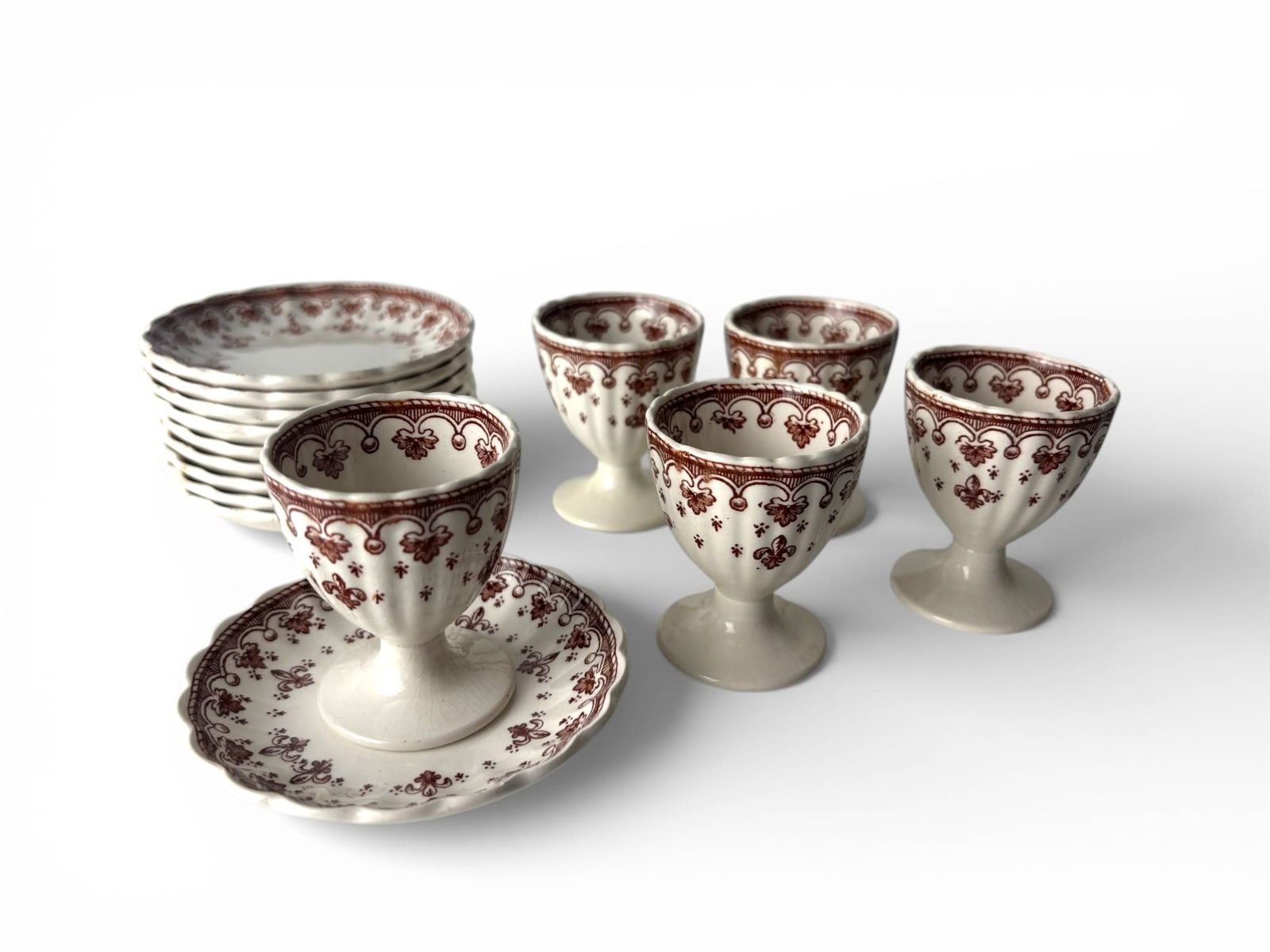 Spode Brown Fleur de Lys Pattern Egg Cups and Saucers: 10 ceramic egg cups with matching saucers by Spode Copeland, England, in the Fleur de Lys pattern. Features scalloped edges and a red floral motif with fleur de lis details. Marked on the base. The ce