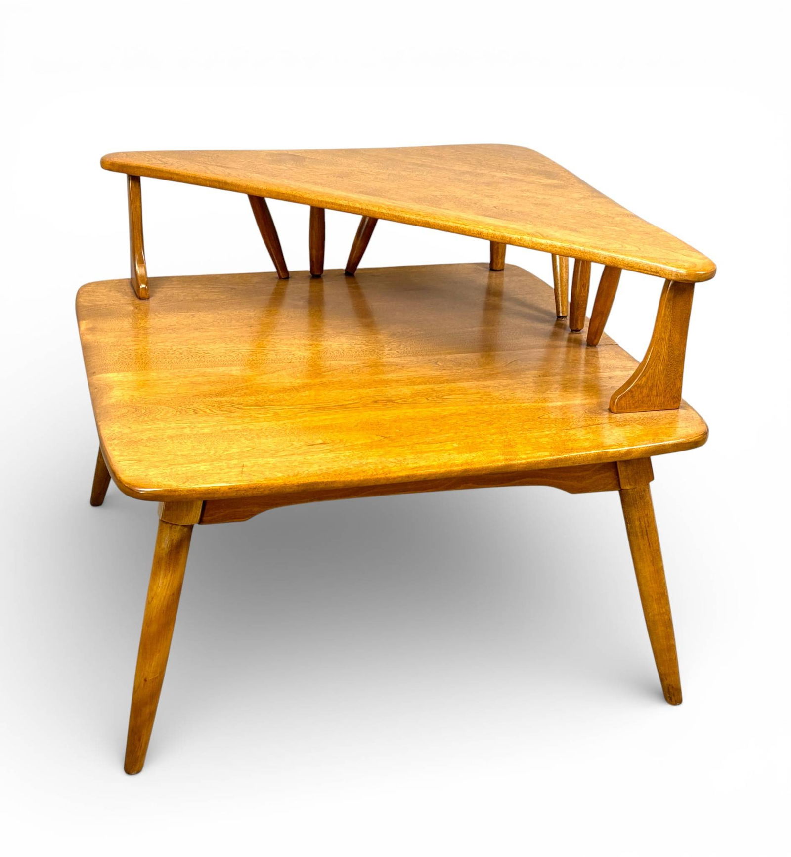 Heywood Wakefield Mid-Century Modern Corner Table in Wheat Finish: Heywood Wakefield Mid Century Modern corner table crafted in solid wood with a two tiered design. Features iconic tapered legs and spindled supports. Finished in the trademark Wheat tone. The seam on