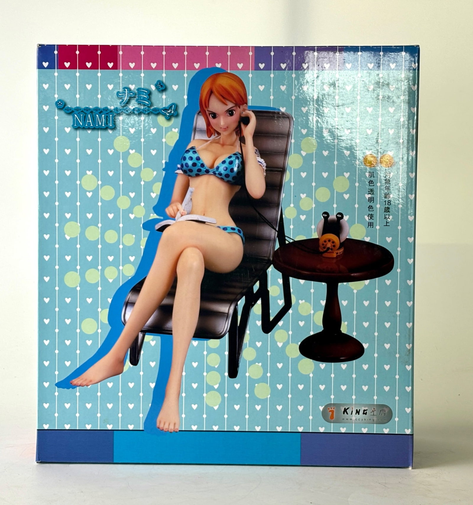 King Studio Nami Polka Dot Bikini Figure in BOX: King Studio figurine featuring the character Nami in a polka dot bikini, seated on a chair with an accessory table. Includes original branded packaging, detailed imagery of Nami, and product markings 