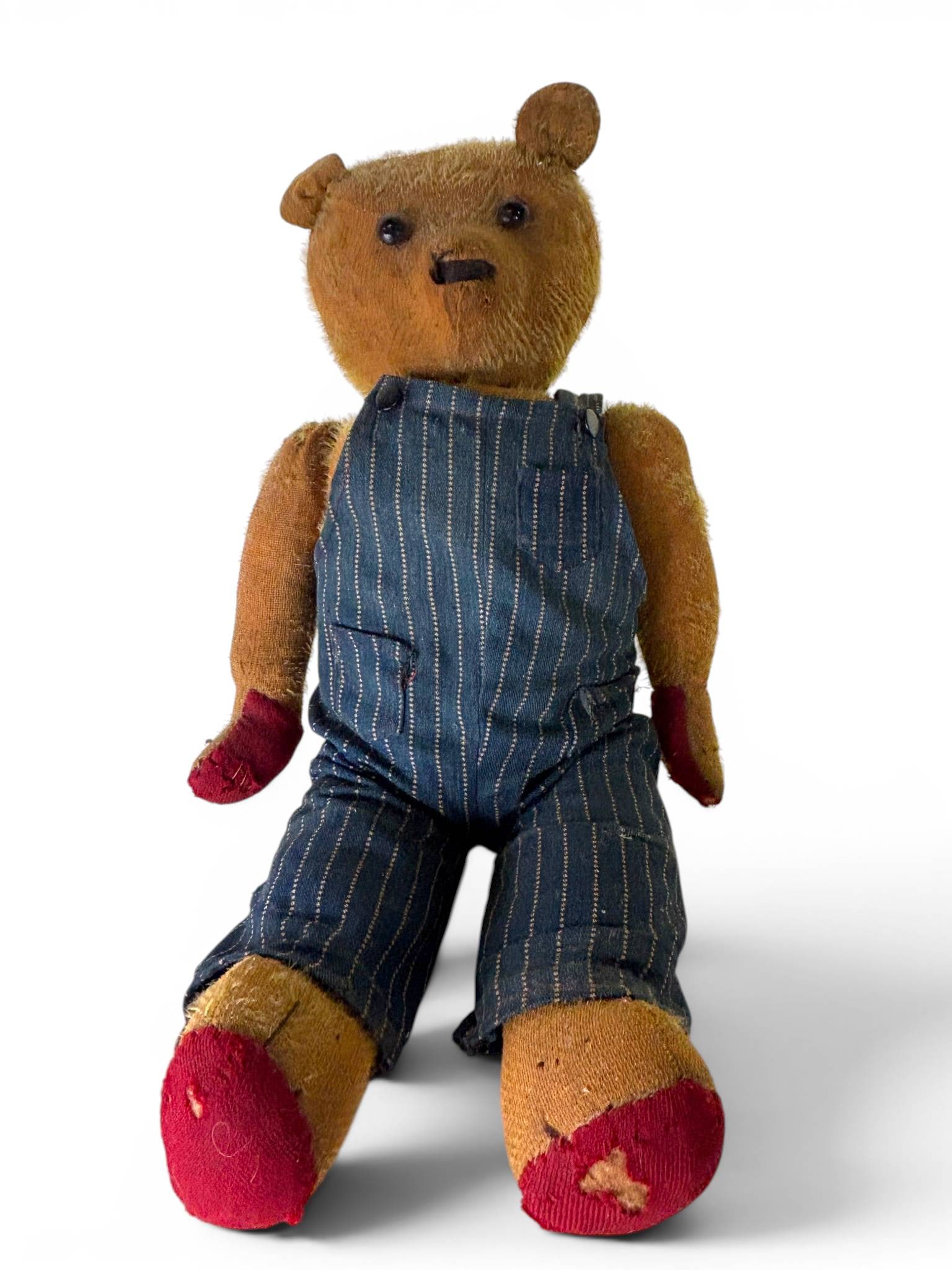 Early Mohair Straw Stuffed Bear in Overalls: An early mohair stuffed teddy bear, featuring straw stuffing and dressed in blue pinstriped overalls. The bear has black button eyes and a stitched nose and mouth. It shows signs of wear, including