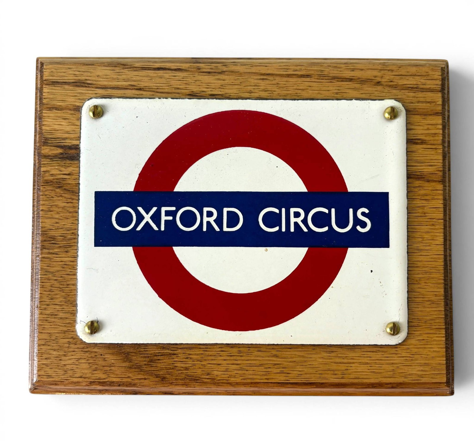 London Underground Oxford Circus Enamel Sign: Enamel sign featuring Oxford Circus in the iconic London Underground roundel design. Sign is mounted on a finished wooden plaque with keyhole slots for wall hanging. The enamel plate is affixed with b