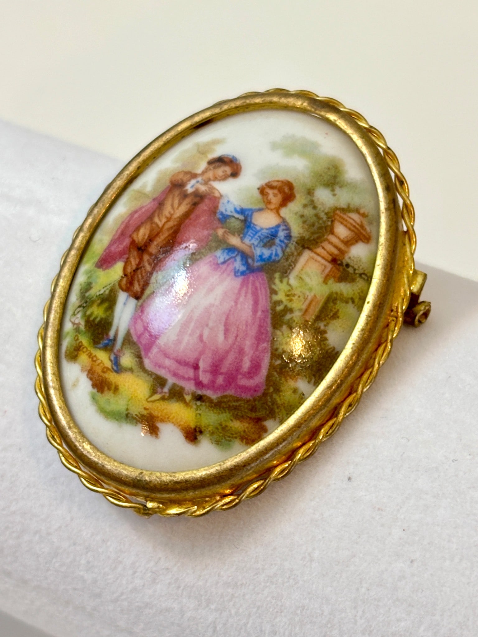 Limoges France Hand Painted Porcelain Brooch: Oval porcelain brooch marked ‘Limoges France’ on the back, featuring a hand painted pastoral scene of a man and woman in period clothing. Encased in a gold tone metal frame with a twisted rope edg