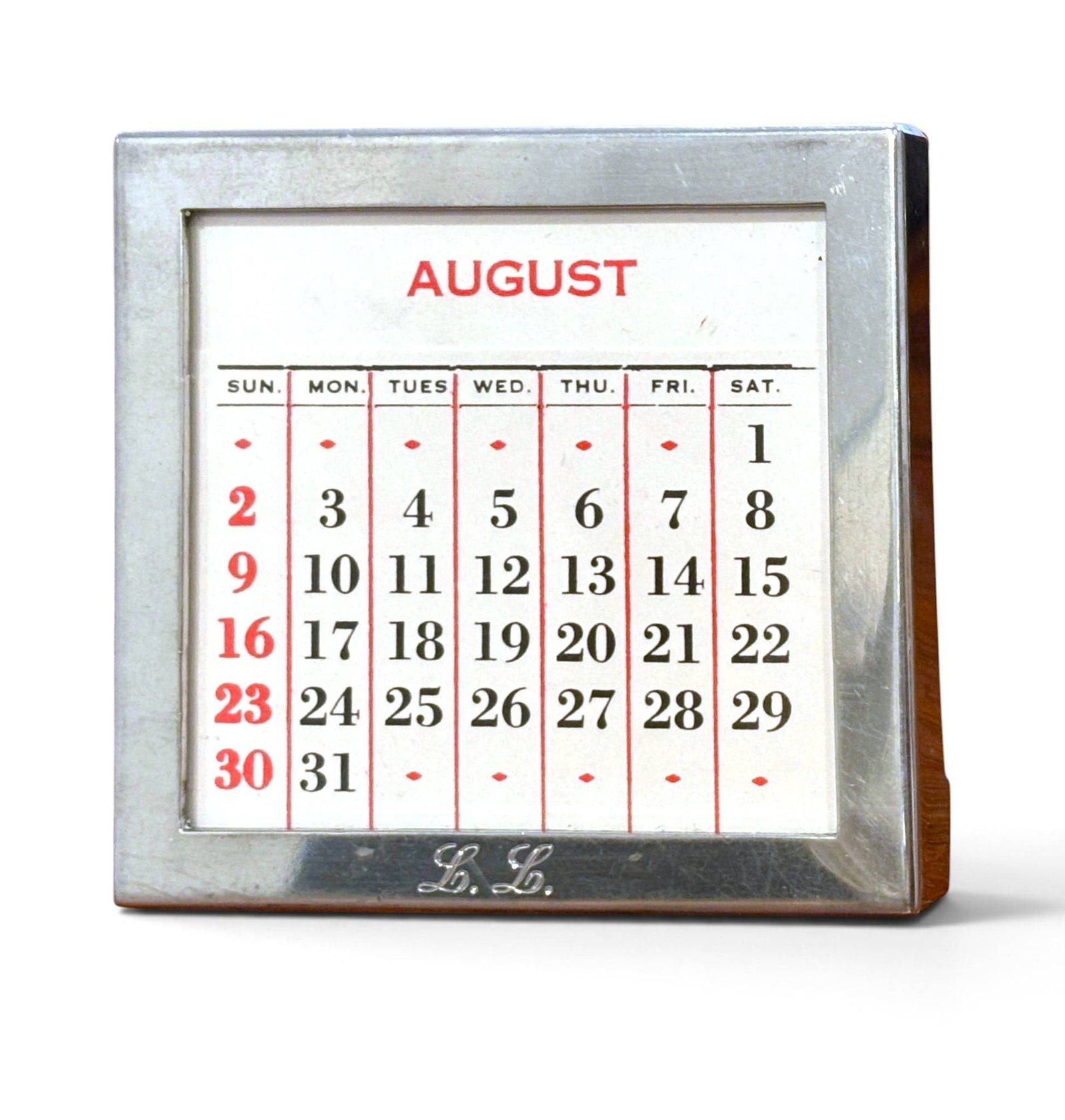 Schroth Pewter Perpetual Calendar, Model 205: Schroth Pewter model 205 perpetual calendar featuring a polished pewter frame with engraved ‘L.L.’ initials on the front. The backing is faux wood with a fold-out stand. Brass corner clips secure 