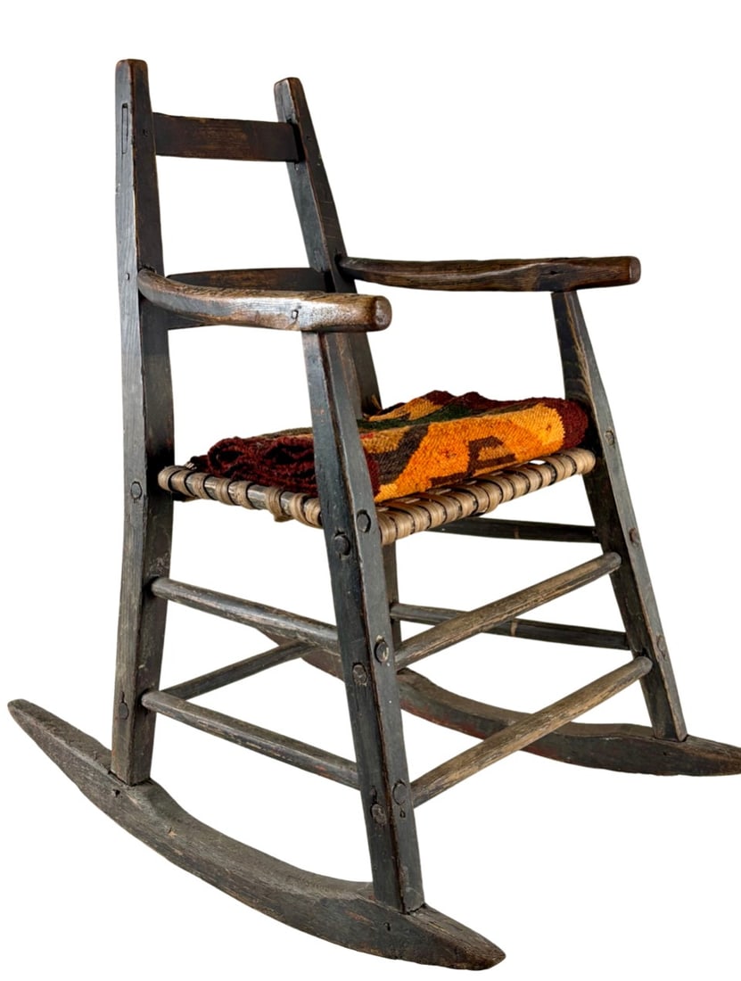 Early American Childs Chair with Rush Seat and Navajo Textile: Early American child’s rocking chair crafted from wood with a woven rush seat. The handmade chair features ladder back support and curved runners. Accompanied by a Navajo textile display, with vibra
