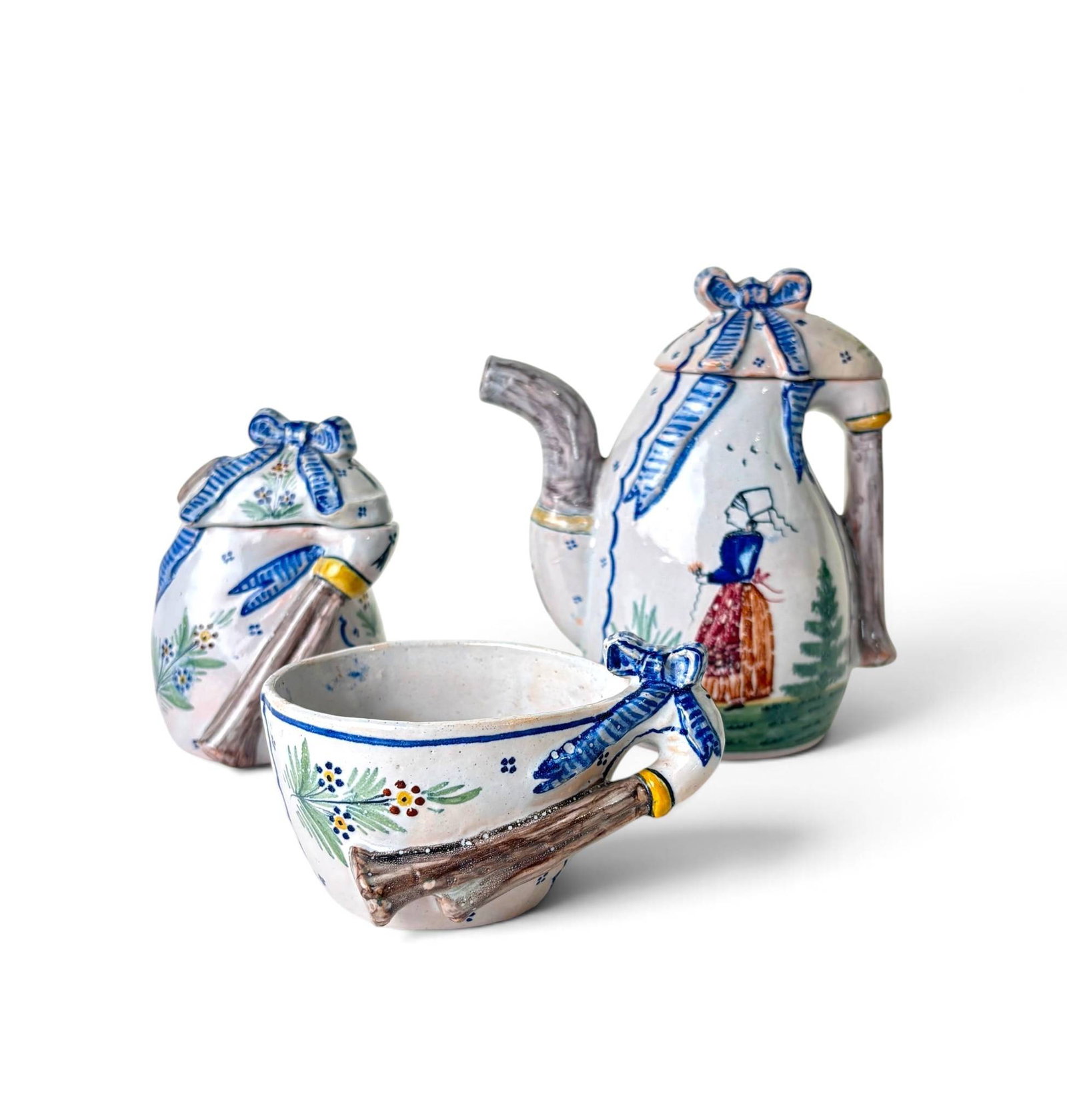 HB Quimper Tea Set Circa 1900-1905: Three ceramic pieces by HB Quimper, including a tea pot, creamer, and cup, hand-painted with blue, green, yellow, and brown details. Designs feature Breton figures, floral motifs, and ribbon accents.