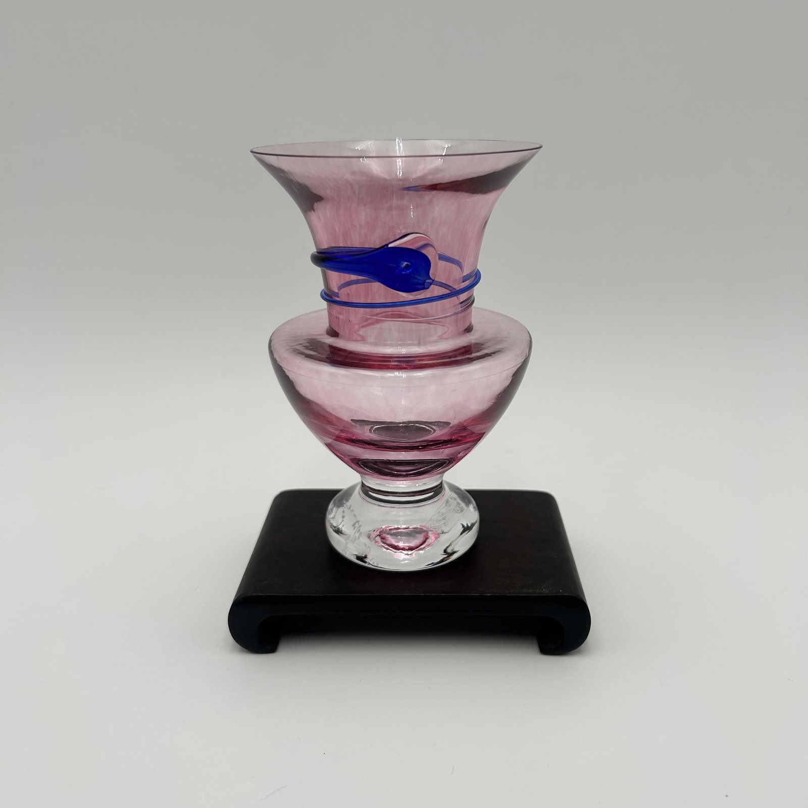 Swedish Kosta Boda "Serpent" Vase by Monica Backstroem: Monica Backstroem (1939-2020) designed many classics for the Swedish Kosta Boda glasswords. This pink vase features a distinctive blue snake motif wrappend around the glass. Comes with Wooden Base. 
