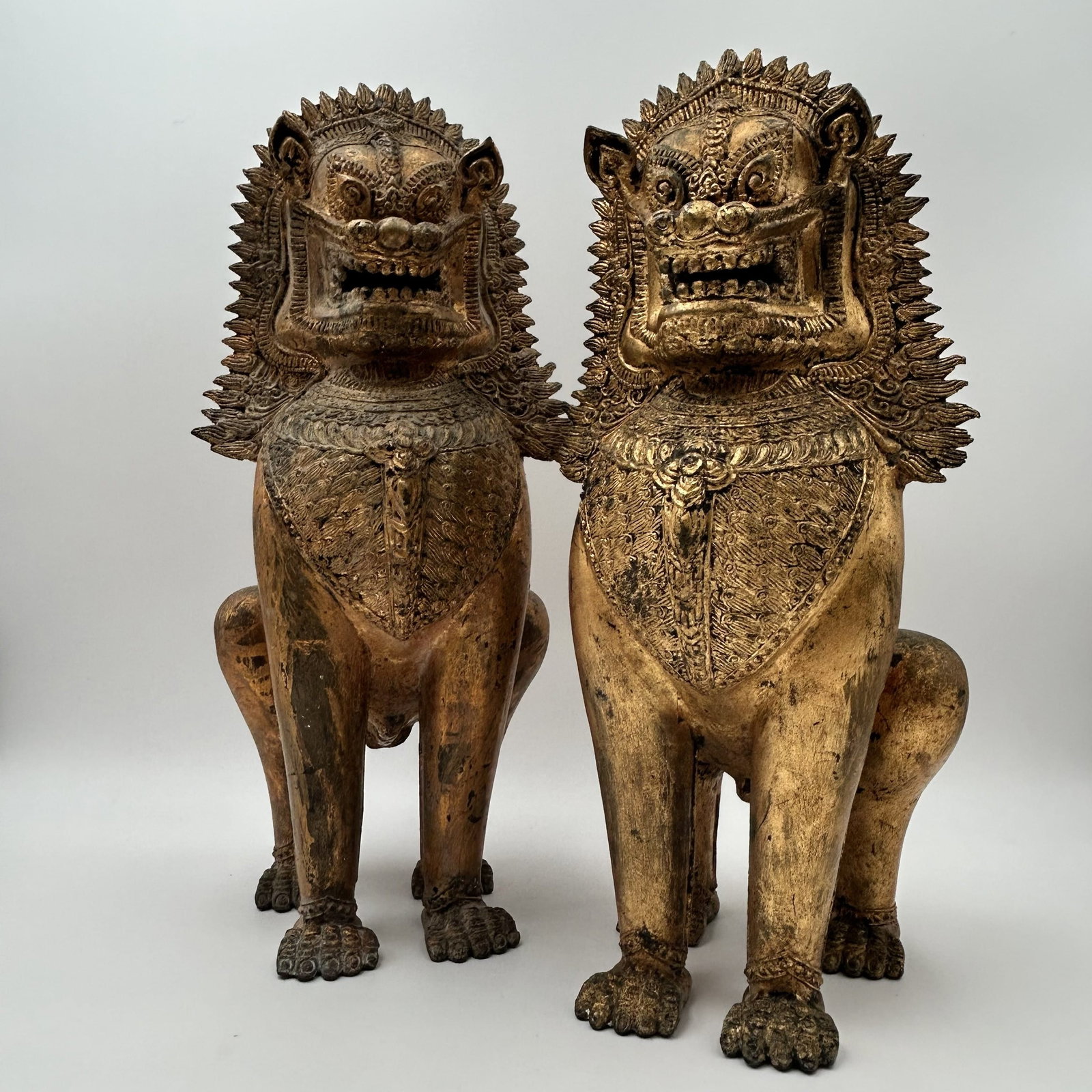 Pair of Thai Gilt Bronze Singha Temple Lions: These are 19th century, cast gilt bronze temple or Buddhist guardian lions having and elaborate collar and headdress. Gilt paint is consistent with age and use. Dimensions: - 7.5" W x 8" D x 17" H eac