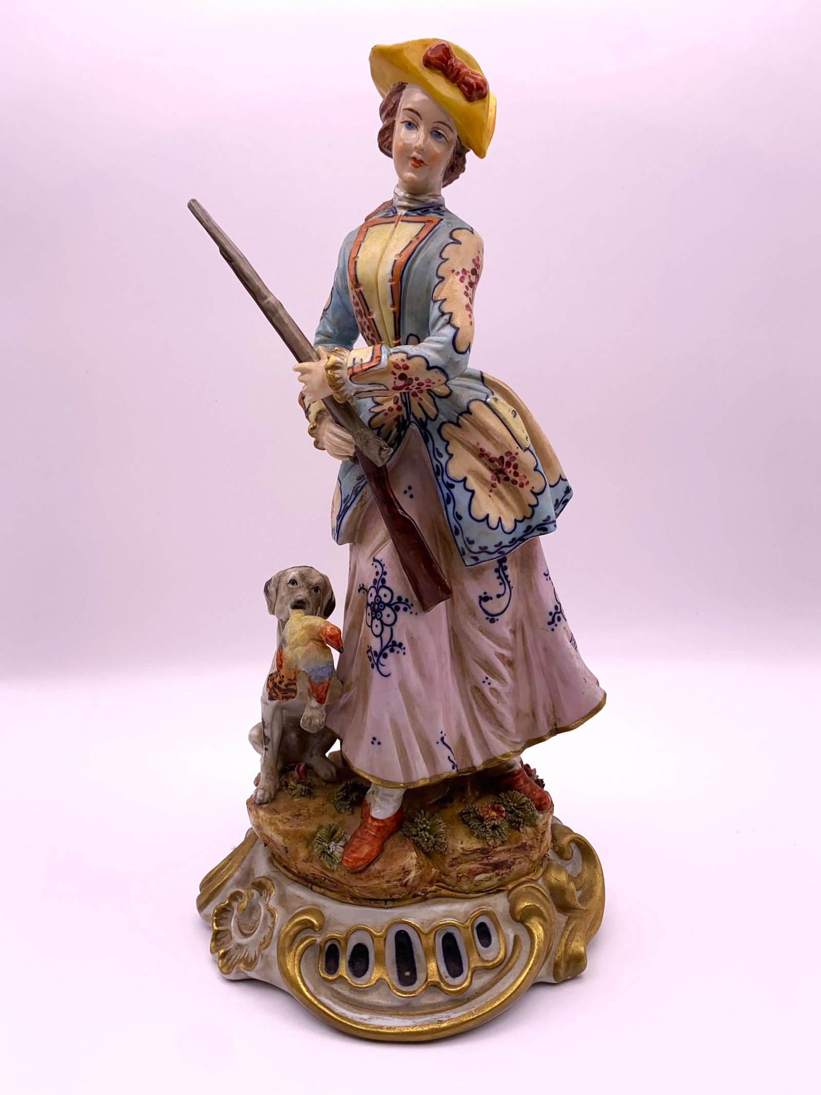 Meissen Statuette of Woman with Hunting Rifle & Dog with Pheasant: Antique statuette from Meissen Porcelain depicting a woman wearing a blue dress and yellow hat holding a hunting rifle, standing next to a hound with a pheasant in its mouth. Meissen crossed swords ma