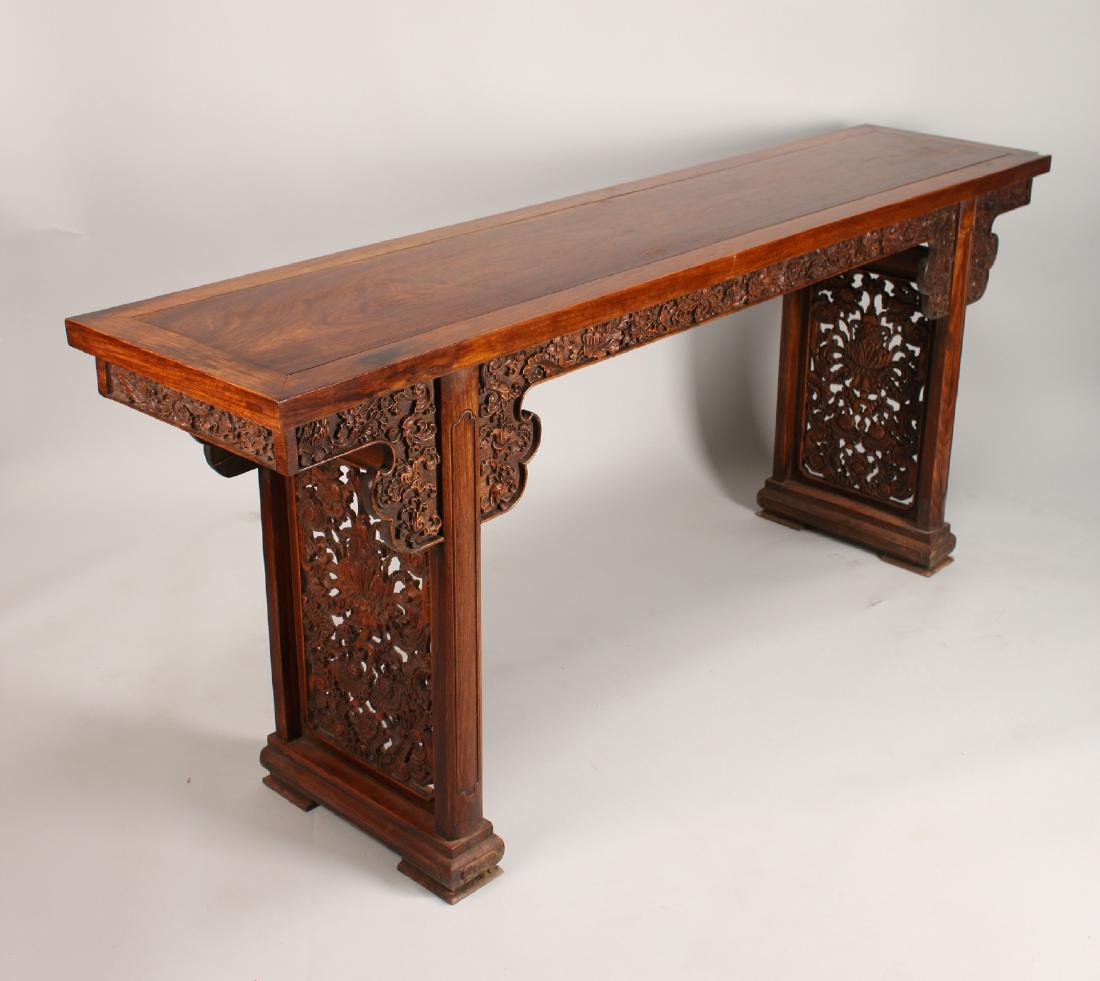 HUANGHUALI PAINTING TABLE: Huanghuali Painting Table: A long Huanghuali painting table with elaborately carved apron and legs. The apron is carved to show lotuses, other flowers, and swirling tendrils. The legs are carved and p