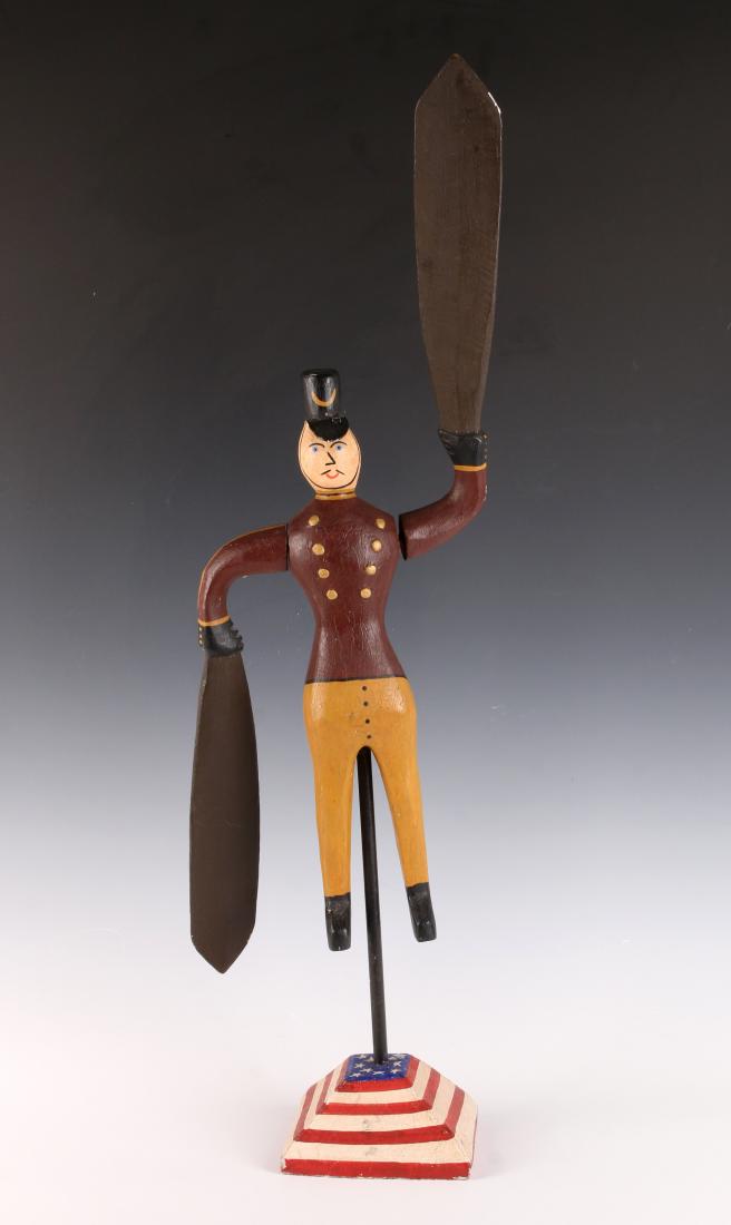 WOODEN FOLK ART WHIRLIGIG: WOODEN FOLK ART WHIRLIGIG : Standing on a triangular base resembling the American flag. His hands are holding the blades that spin. Dressed in gold pants and a double breasted burgundy jacket. Wearing