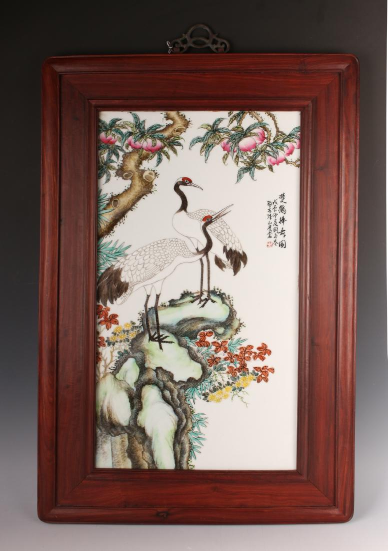 PLAQUE OF CRANES: PLAQUE OF CRANES: Pair of cranes are under a peach tree and standing on a boulder with flowers growing on the underside. Calligraphy and artist marks on upper right. In a wide wooden frame with a bron