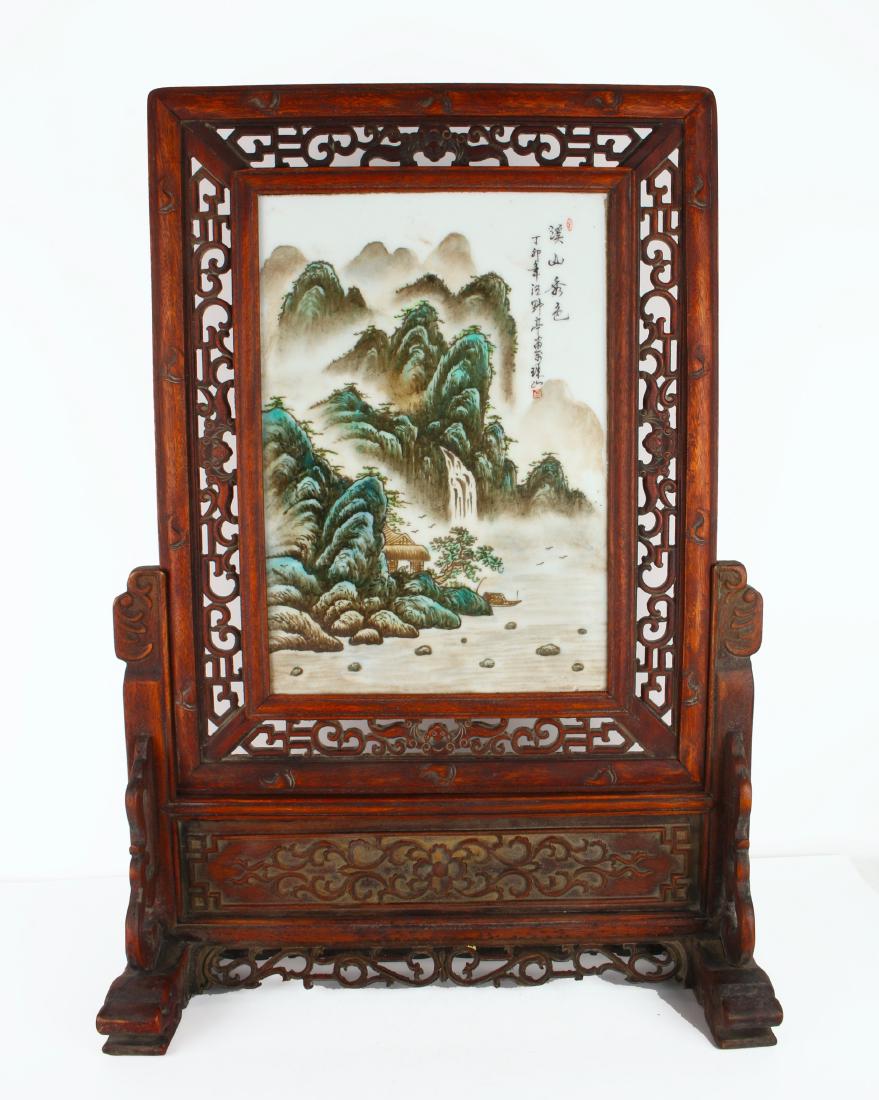 TABLE SCREEN WITH PORCELAIN PLAQUE OF MOUNTAINS: TABLE SCREEN WITH PORCELAIN PLAQUE OF MOUNTAINS : A waterfall cascades into a river that flows through the bottom right of the scene. A rocky riverbank sits to the left. Misty mountains rise in the di