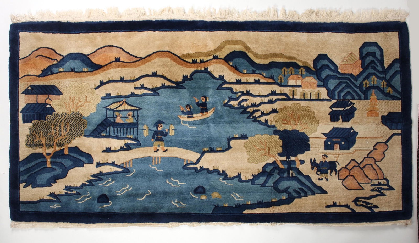 1920'S CHINESE SILK RUG: 1920'S CHINESE SILK RUG: A landscape scene in blues and beiges. There are two people in a boat in the water with a man carrying his goods across a bridge. Mountains, trees, and boulders accent the are